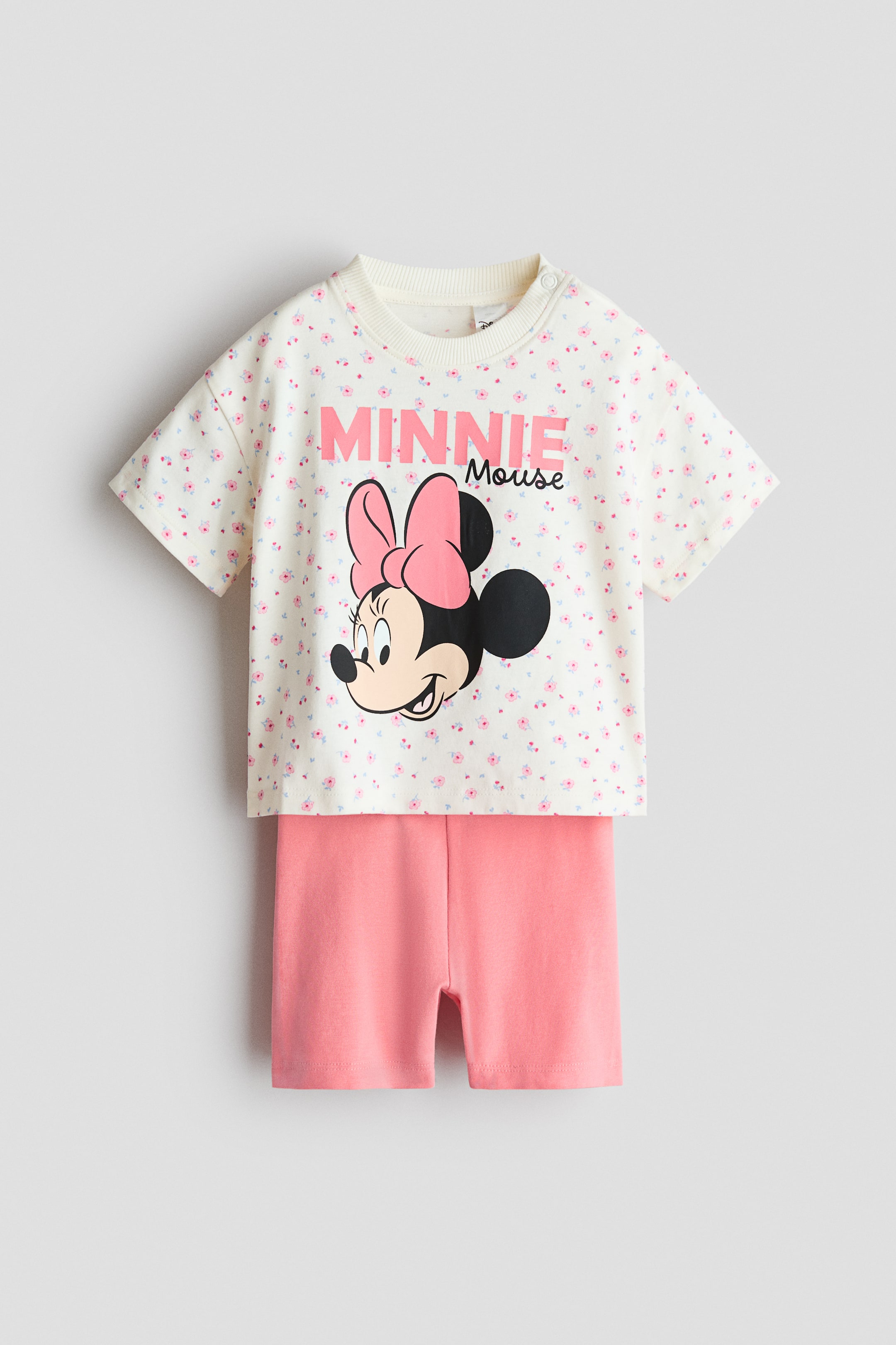 Ver imagen más grande: Se presenta un conjunto de ropa infantil de dos piezas, visto de frente, que incluye una camiseta de manga corta en color blanco roto con un estampado floral rosa, un cuello redondo acanalado con un botón de presión en el hombro izquierdo, y un gran gráfico que muestra la cabeza de Minnie Mouse y el texto "MINNIE Mouse". Se combina con unos pantalones cortos de color rosa claro sólido.