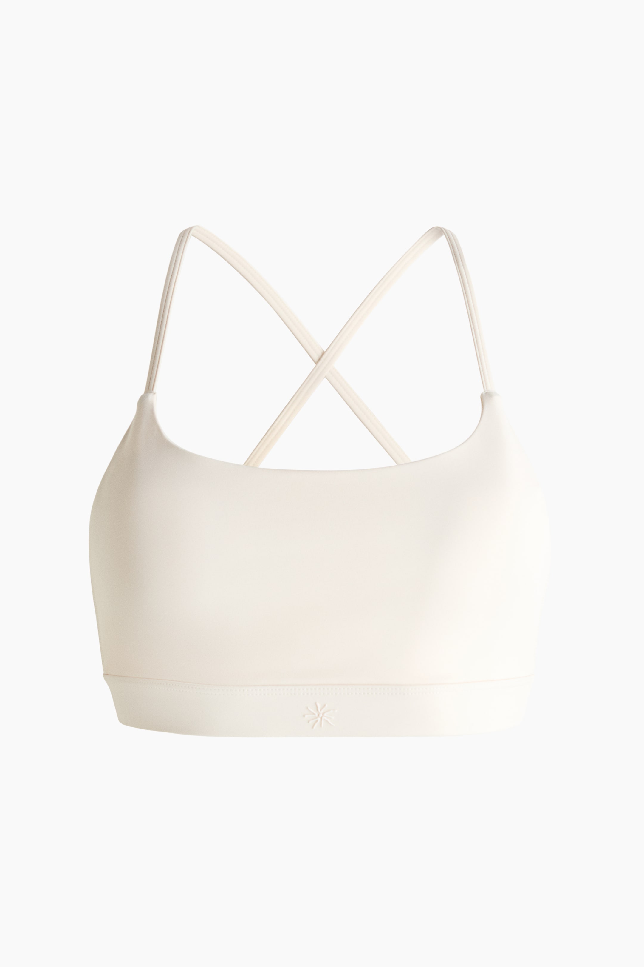 Ver imagem maior: Um sutiã desportivo em branco sujo é apresentado de frente, com um decote em U e alças finas que formam um design cruzado. Uma faixa elástica larga forma a bainha inferior, exibindo um pequeno logótipo em forma de estrela no lado esquerdo.