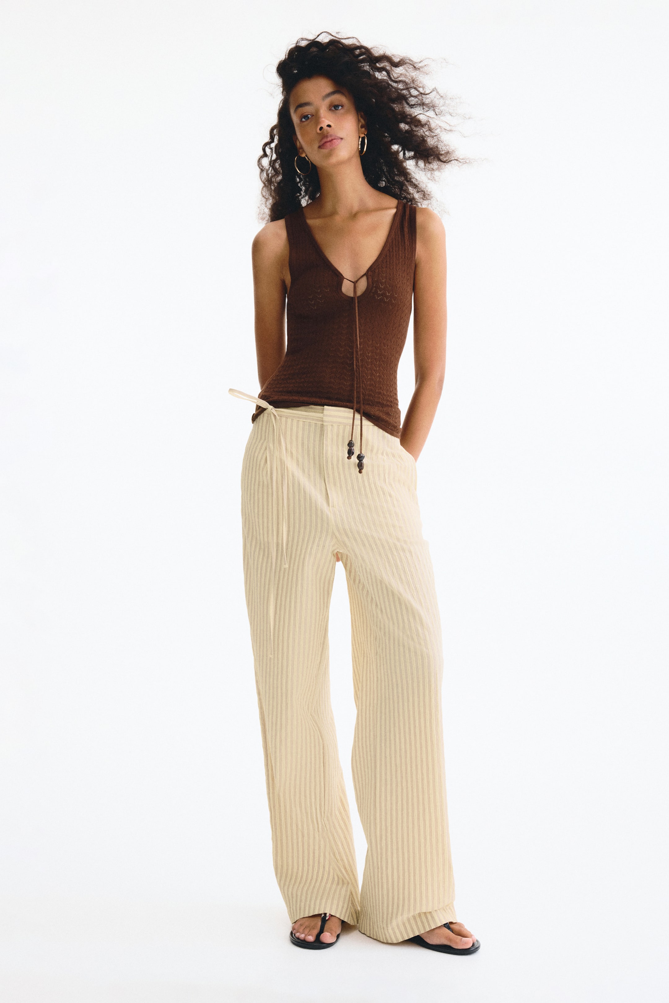View larger image: The model is wearing light beige woven pants with vertical stripes. These pants have a regular waist and a narrow tie detail at the front. The straight legs feature pleats at the top, and there are diagonal side pockets where the model is placing their hands.