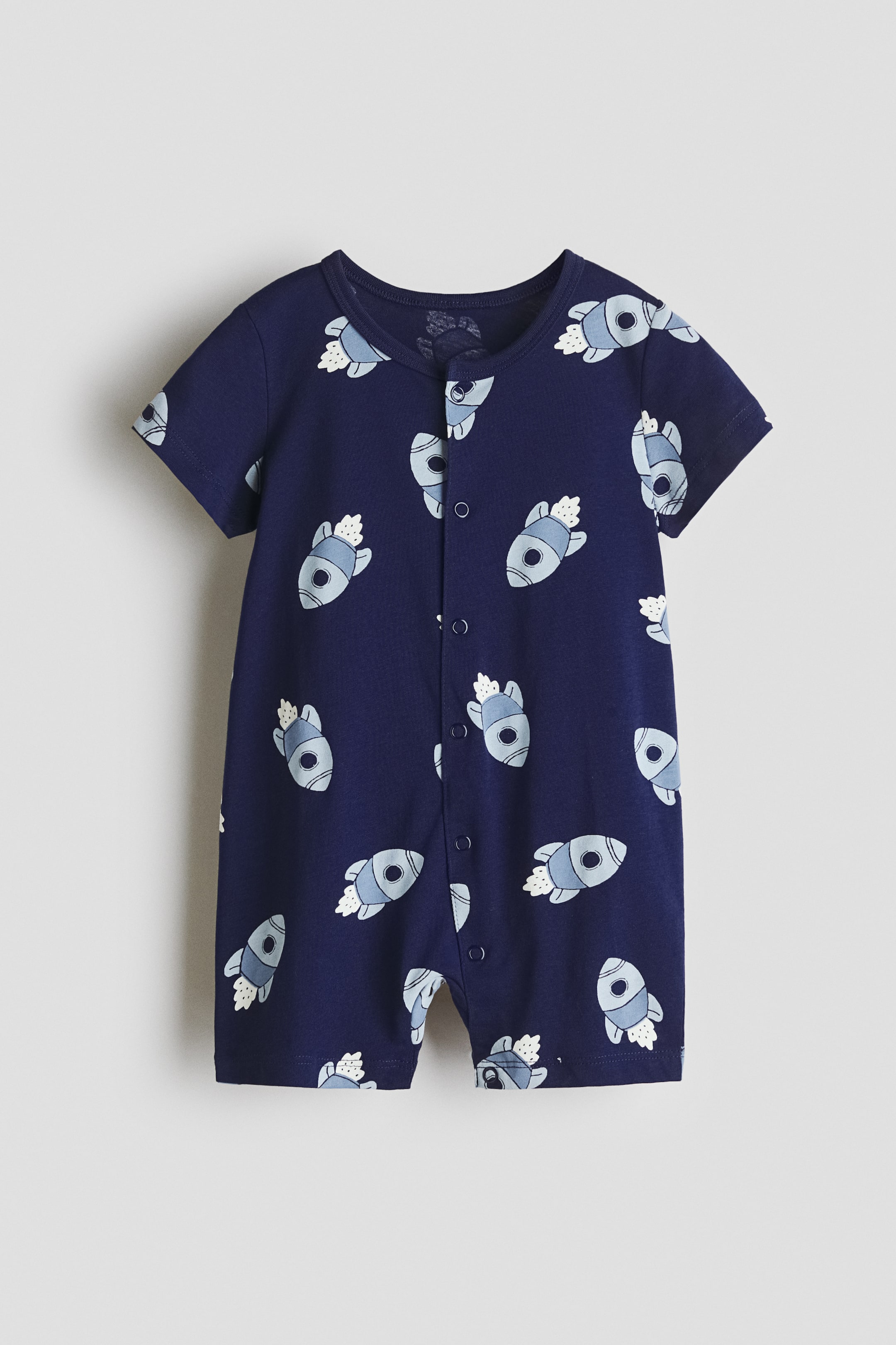 View larger image: The front view of a short-leg sleepsuit showcases a navy blue cotton jersey adorned with a repeating light blue and white rocket ship pattern. It features a round neckline, short sleeves, and press-studs running down the front and along one leg.