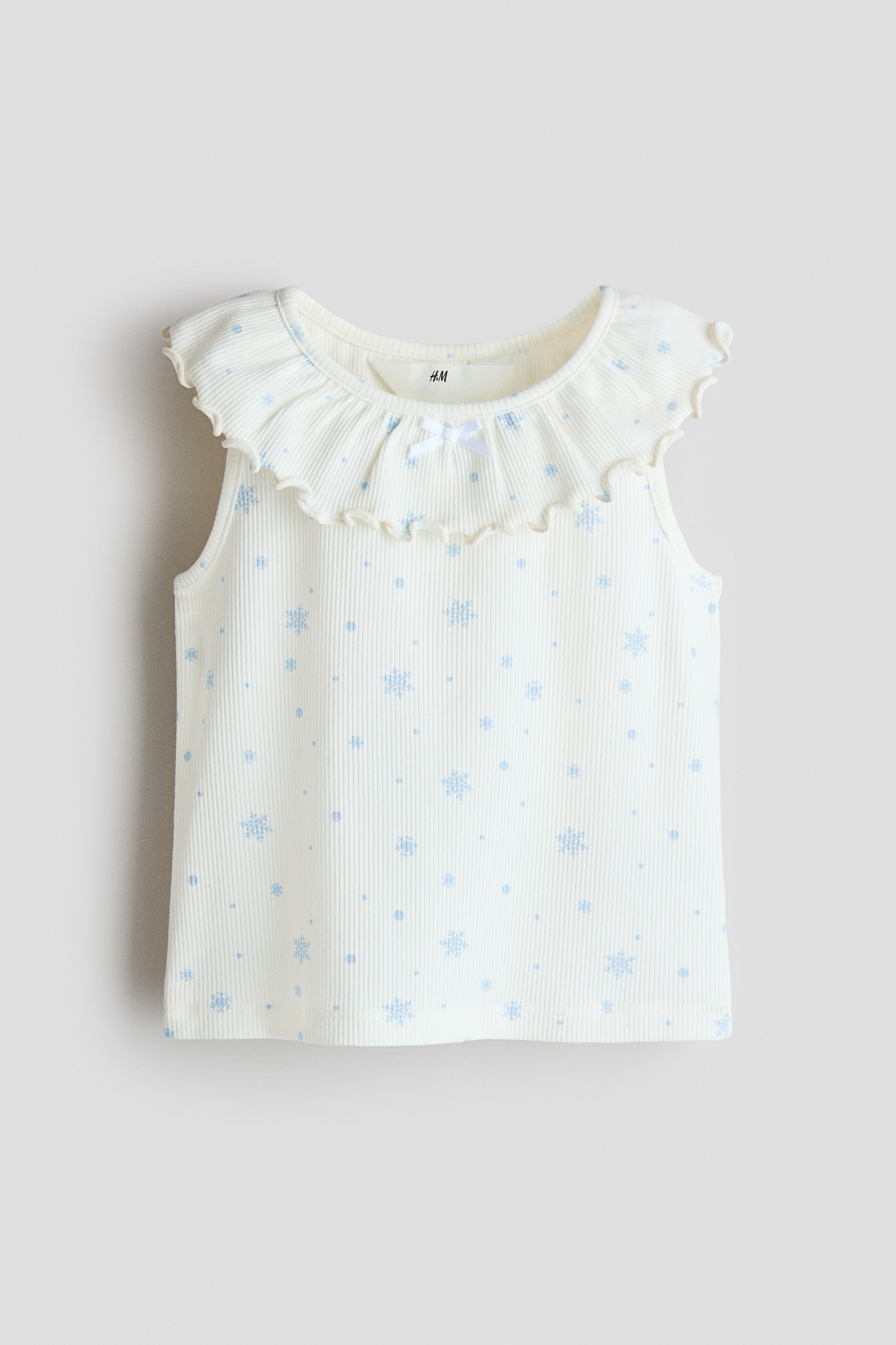 The model is wearing a sleeveless top in off-white ribbed cotton jersey with a blue snowflake print and a ruffled flounce collar. The top is styled with matching ribbed shorts featuring the same print.