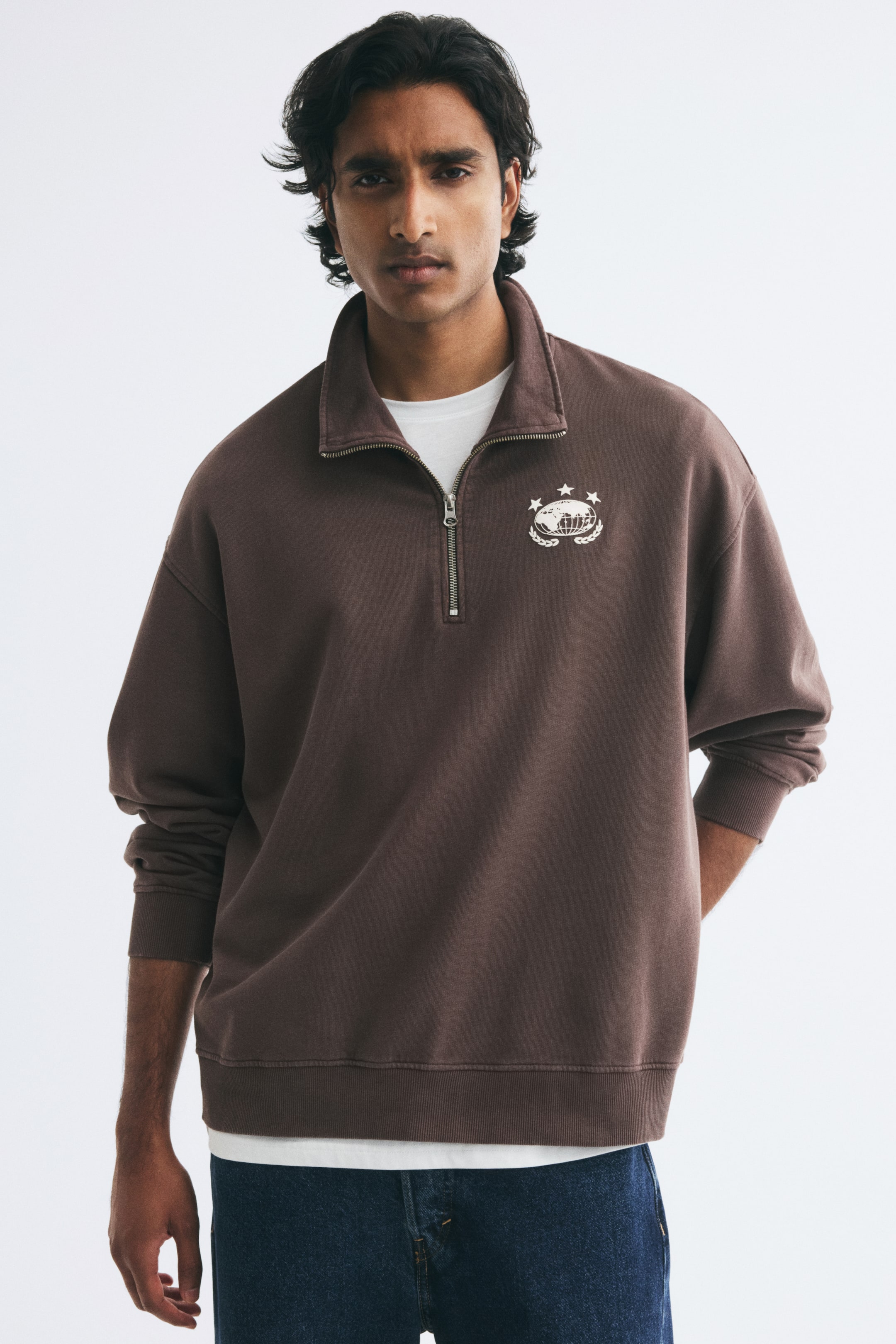 View larger image: The model is wearing a brown quarter-zip sweatshirt with a white globe graphic on the left chest. The sweatshirt is layered over a white T-shirt and styled with blue denim jeans, featuring a stand-up collar with a partially open silver zipper.