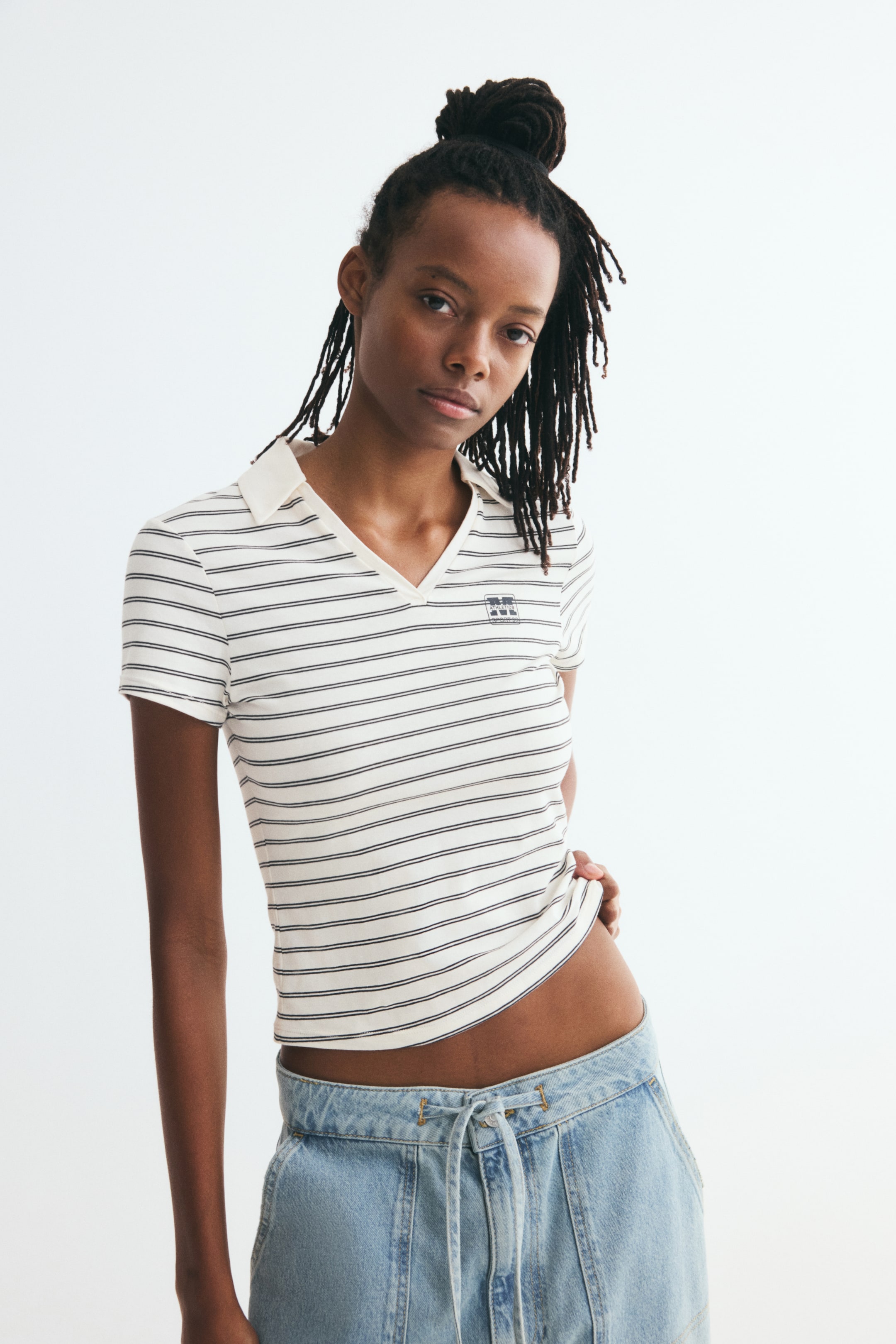 View larger image: The polo shirt is shown from the front, highlighting a cream-colored cotton jersey fabric with thin horizontal black stripes and a small printed logo on the left chest. It features an open V-neck collar, short sleeves, and a straight-cut hem that sits just above the waistline of light-wash denim jeans.