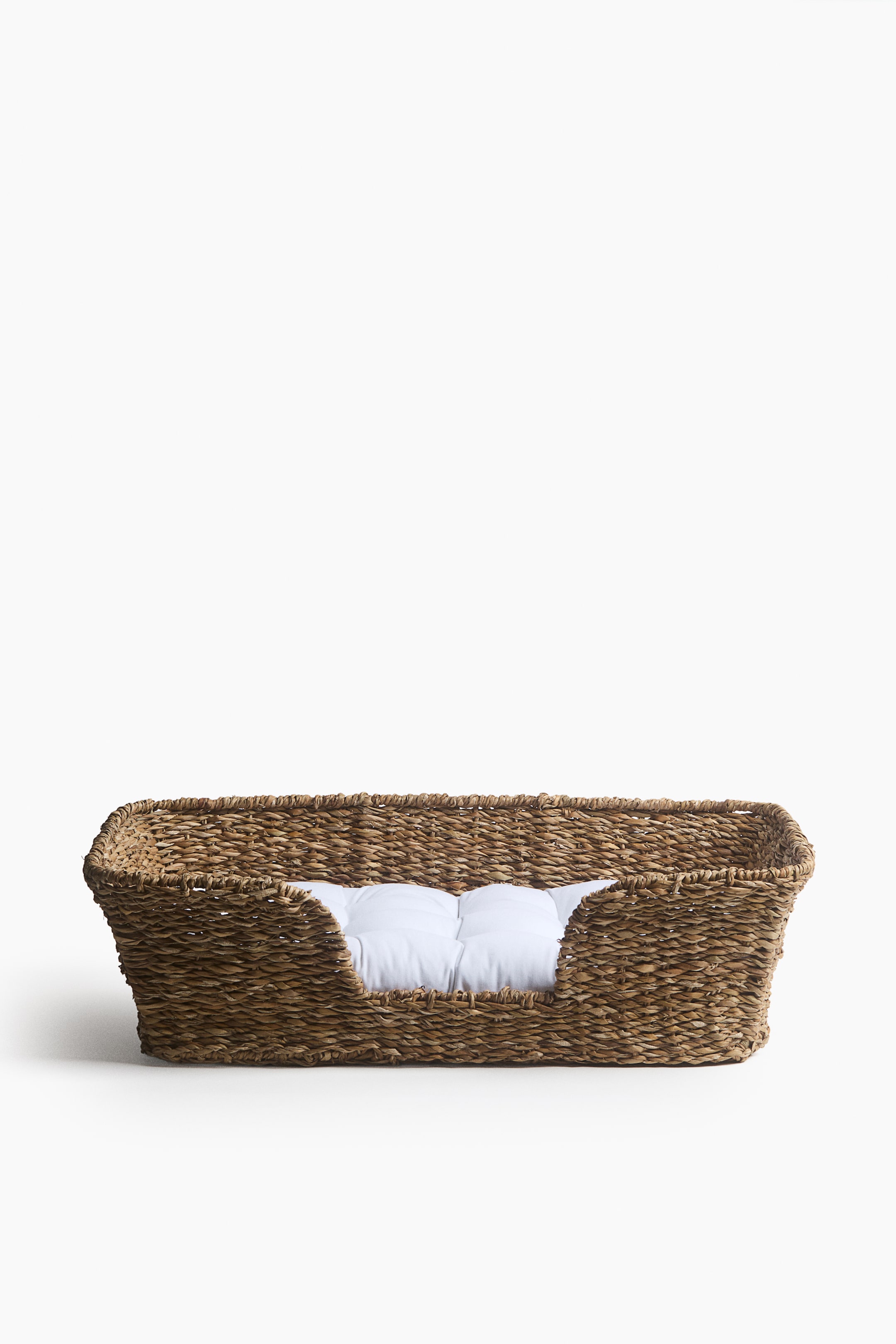 View larger image: A rectangular pet bed woven from natural brown fibres is facing the front, featuring high sides and a lowered front entryway. A white, tufted fabric cushion is resting inside the basket.