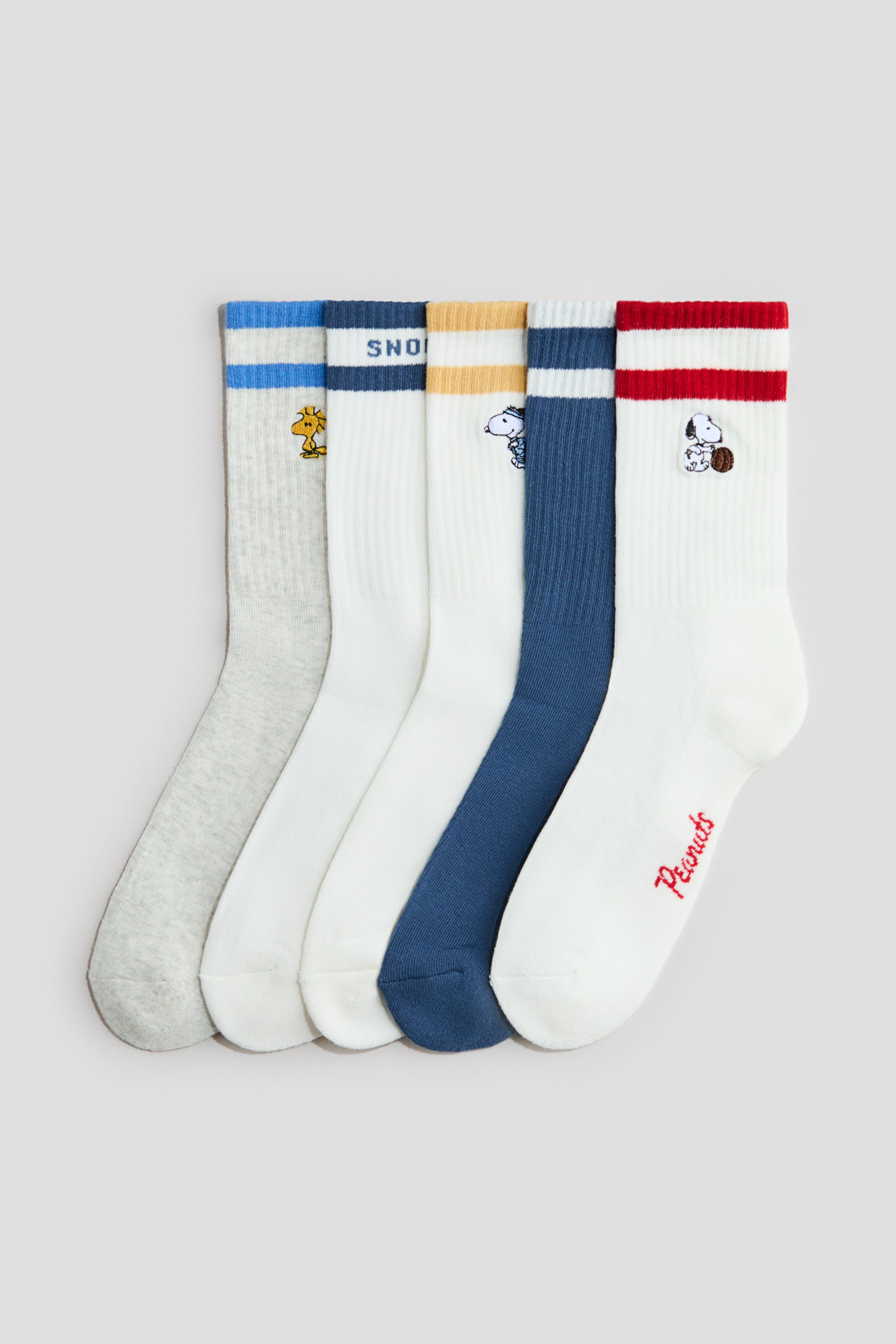 Größeres Bild ansehen: Ein Set aus fünf Feinstricksocken liegt flach, wobei jede Socke einen gerippten Schaft und einen elastischen Abschluss am oberen Rand aufweist. Jede Socke zeigt ein Intarsienmotiv eines Peanuts-Charakters oder Textes, zusammen mit gestreiften Bündchen in verschiedenen Farbkombinationen.