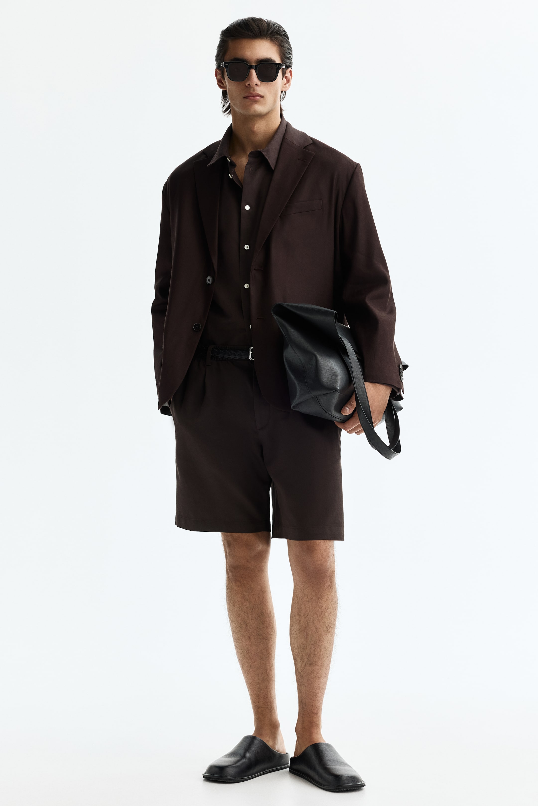 Ingrandisci l'immagine: Un outfit monocromatico marrone scuro viene presentato, caratterizzato da un blazer aperto a vestibilità rilassata con revers a dente sopra una camicia con colletto, abbinato a bermuda al ginocchio e una cintura intrecciata nera. Occhiali da sole con montatura quadrata nera riposano sul viso, una borsa a secchiello in pelle nera è tenuta in mano e sabot neri completano l'insieme.