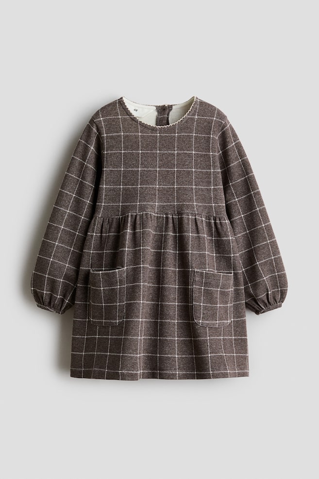 A brown jacquard jersey dress in a white grid pattern is shown from the front, featuring a round neckline with picot trim and long sleeves with elasticated cuffs. The dress has a gathered seam at the waist and a flared skirt with two front patch pockets. - 1