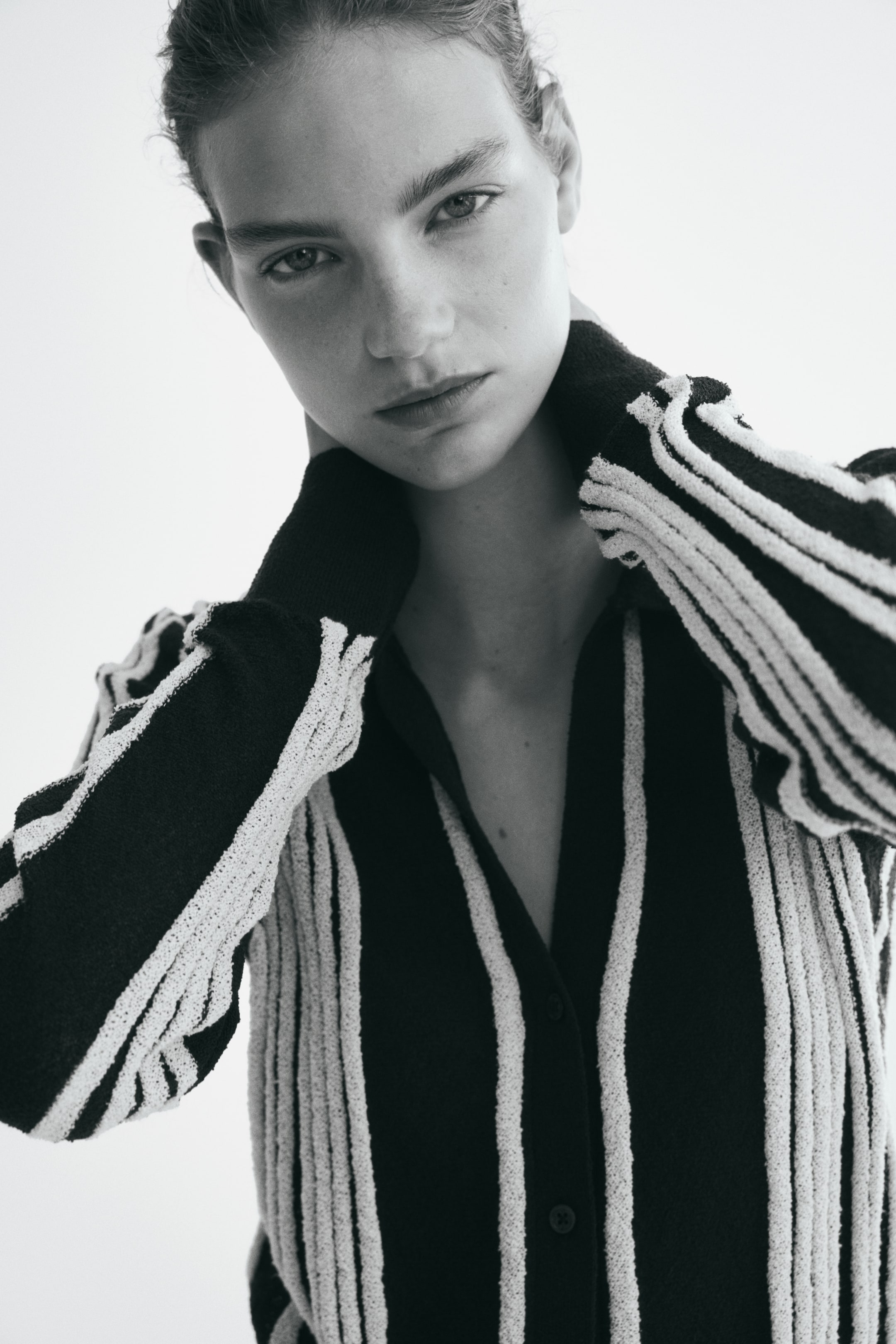 Agrandir l'image: Un cardigan ajusté en maille côtelée irrégulière noire et blanche à rayures verticales, avec un col ouvert et une fermeture boutonnée sur le devant, est porté par le mannequin. Le mannequin pose les deux mains contre l’arrière de son cou, mettant en valeur les manches longues du cardigan ainsi que ses poignets noirs unis.