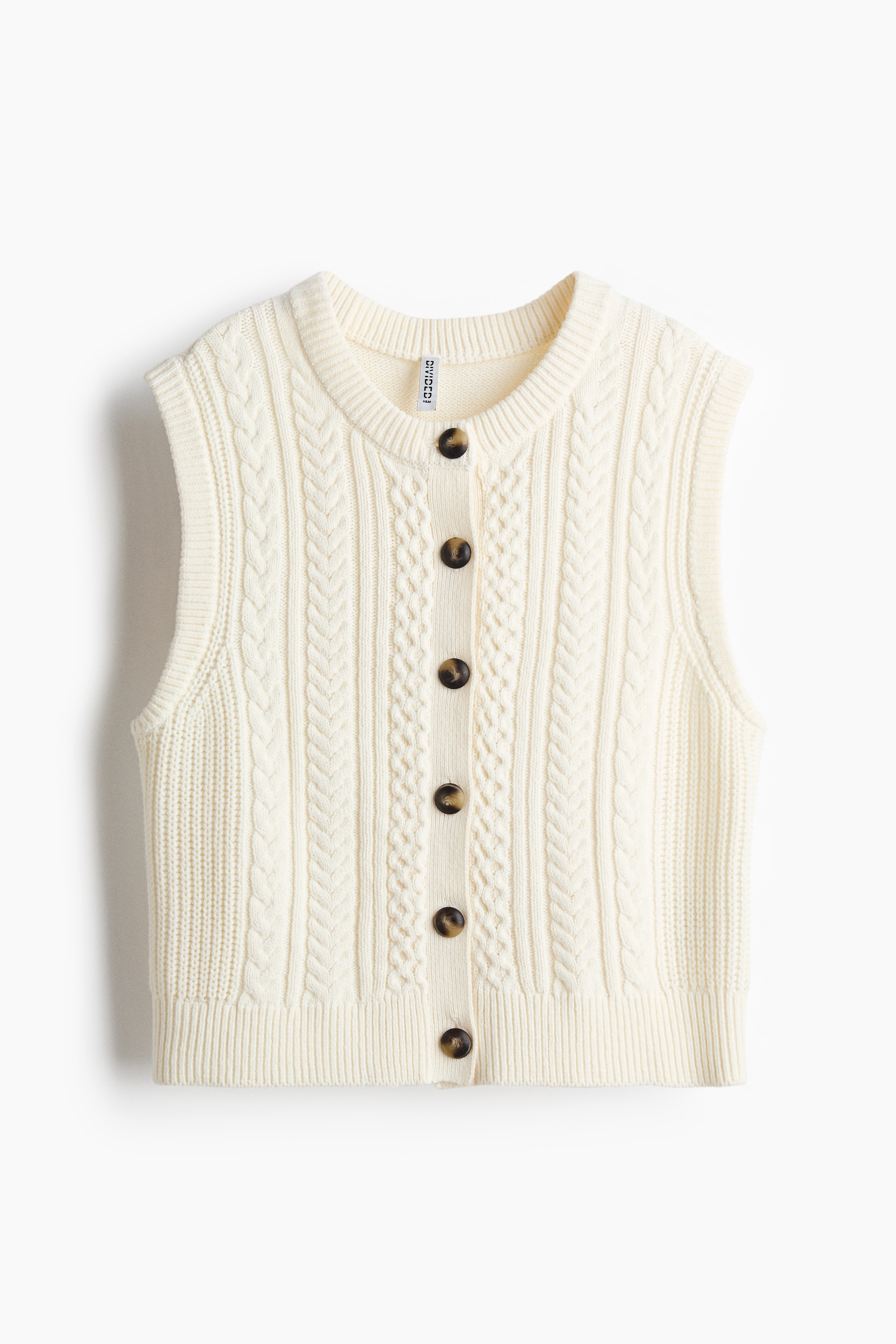 A short, cream-coloured knit vest is shown from the front, featuring a cable knit pattern, a round ribbed neckline, and dark buttons down the front. The sleeveless vest is displaying ribbed trim along the armholes and hem, and is styled with wide-leg brown trousers.