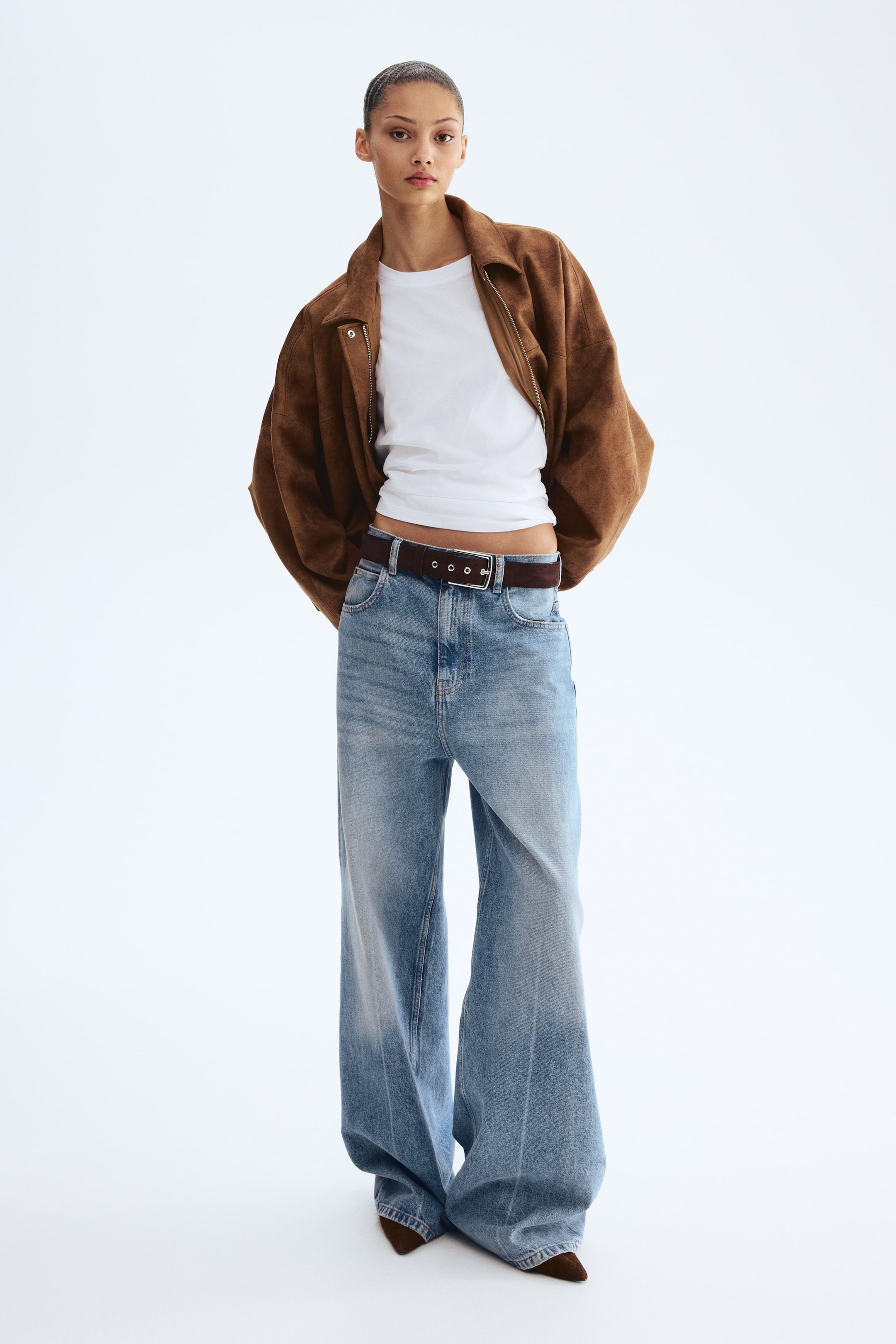 View larger image: A stylish ensemble showcases a brown suede-like oversized jacket, worn open over a fitted white crew-neck top. Light blue wide-leg jeans are cinched by a dark brown belt, with brown pointed-toe shoes peeking out at the bottom.