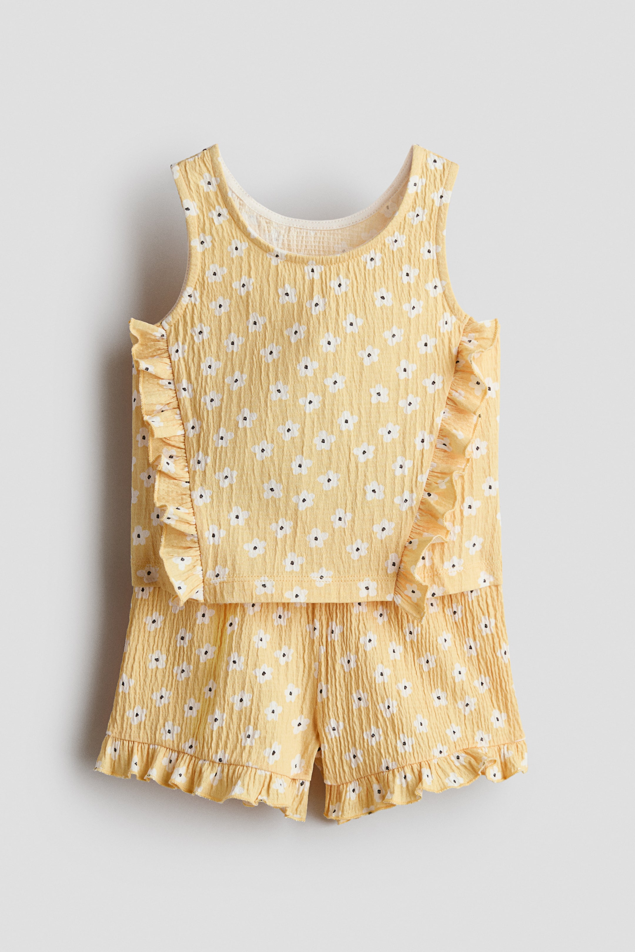 Grotere afbeelding bekijken: Een set met een hemdje en shorts in geel gekreukte jersey is van voren te zien, met een witte bloemenprint. Het hemdje is ontworpen met een ronde halslijn en ruches die langs de voorkant lopen, terwijl de shorts een elastische tailleband en ruches aan de zomen hebben.