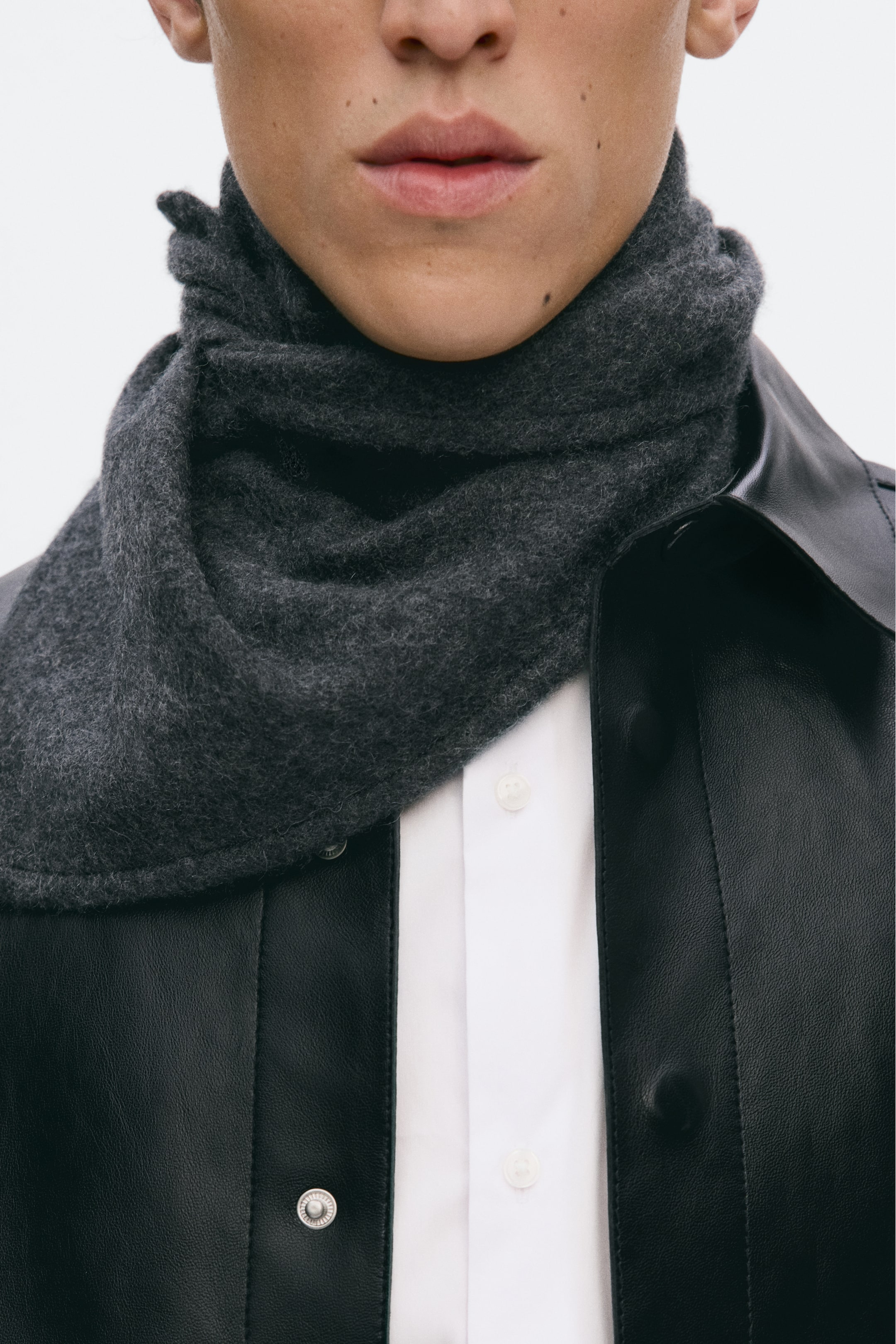 Größeres Bild anzeigen: Ein dunkelgraues Dreieckstuch aus weichem, gefilztem Kaschmir wird eng um den Hals des Models gewickelt, mit einem sichtbaren Knoten am Rücken. Es wird über einem weißen Hemd und unter einer schwarzen Jacke getragen.