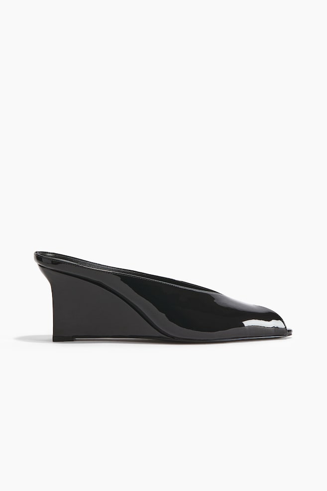 Une mule en cuir verni noir à talon compensé est présentée en profil latéral. Elle présente un décolleté bas avec une ouverture en V profond et un bout subtilement carré, tandis que le talon compensé solide s'intègre parfaitement à la semelle. - 1