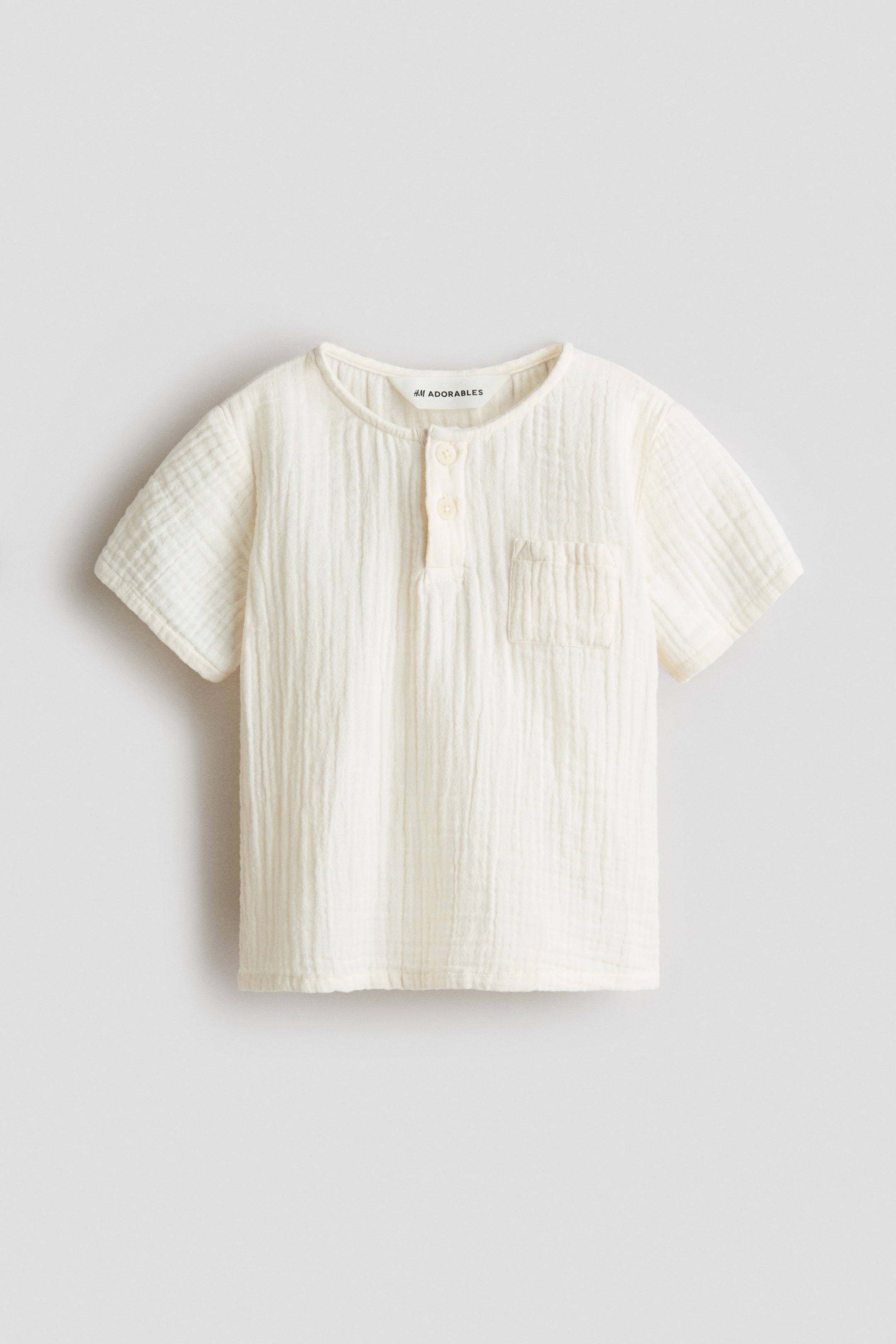 View larger image: An off-white top featuring a muslin texture, a round, narrow-trimmed neckline, a two-button placket, short sleeves, and a patch chest pocket, all shown from the front.