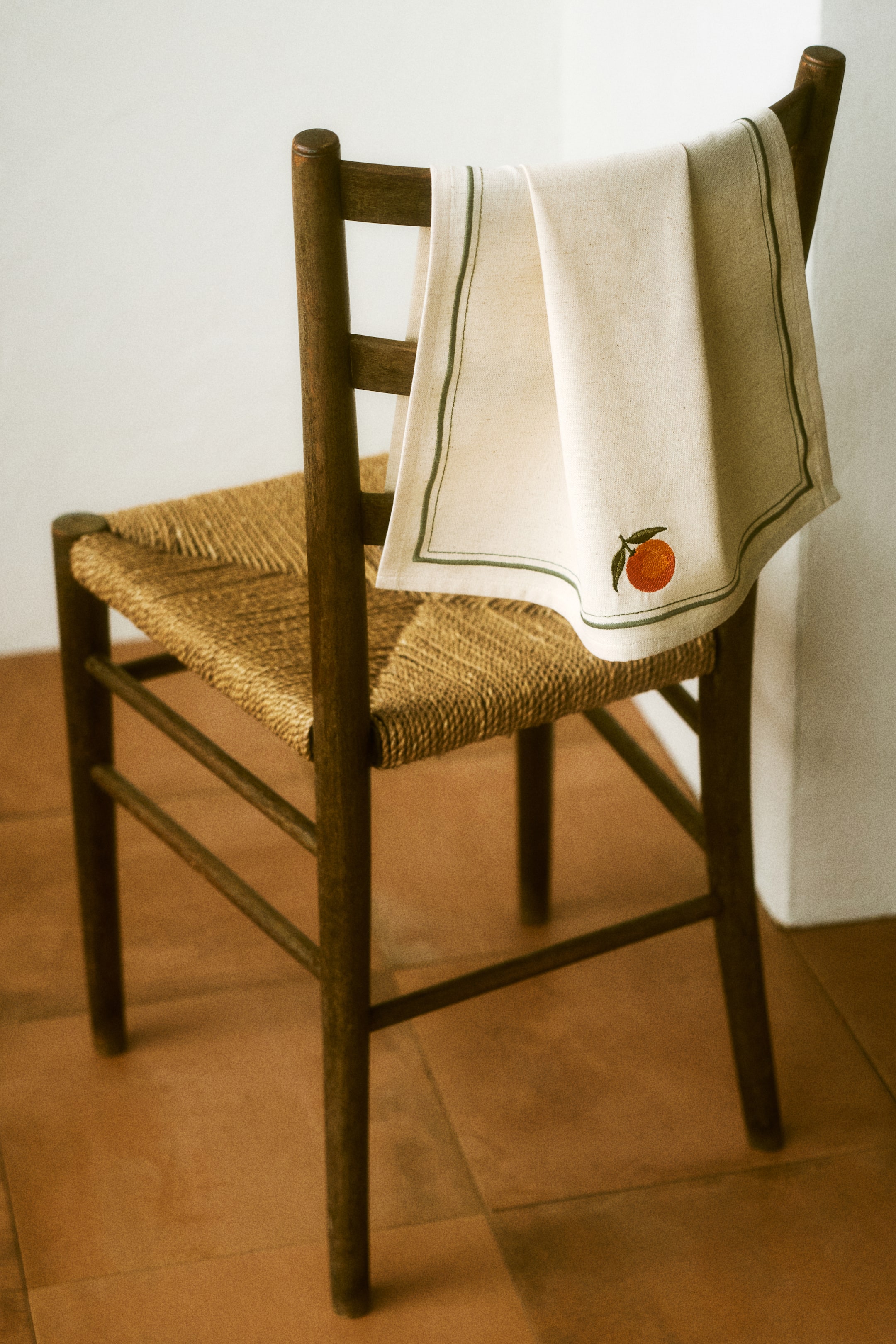 Ampliar la imagen: Un paño de cocina está colgado sobre el respaldo de una silla, mostrando su parte frontal. Está hecho de una mezcla de algodón y lino de color natural, con un delgado borde bordado en verde y una naranja bordada con una hoja verde.
