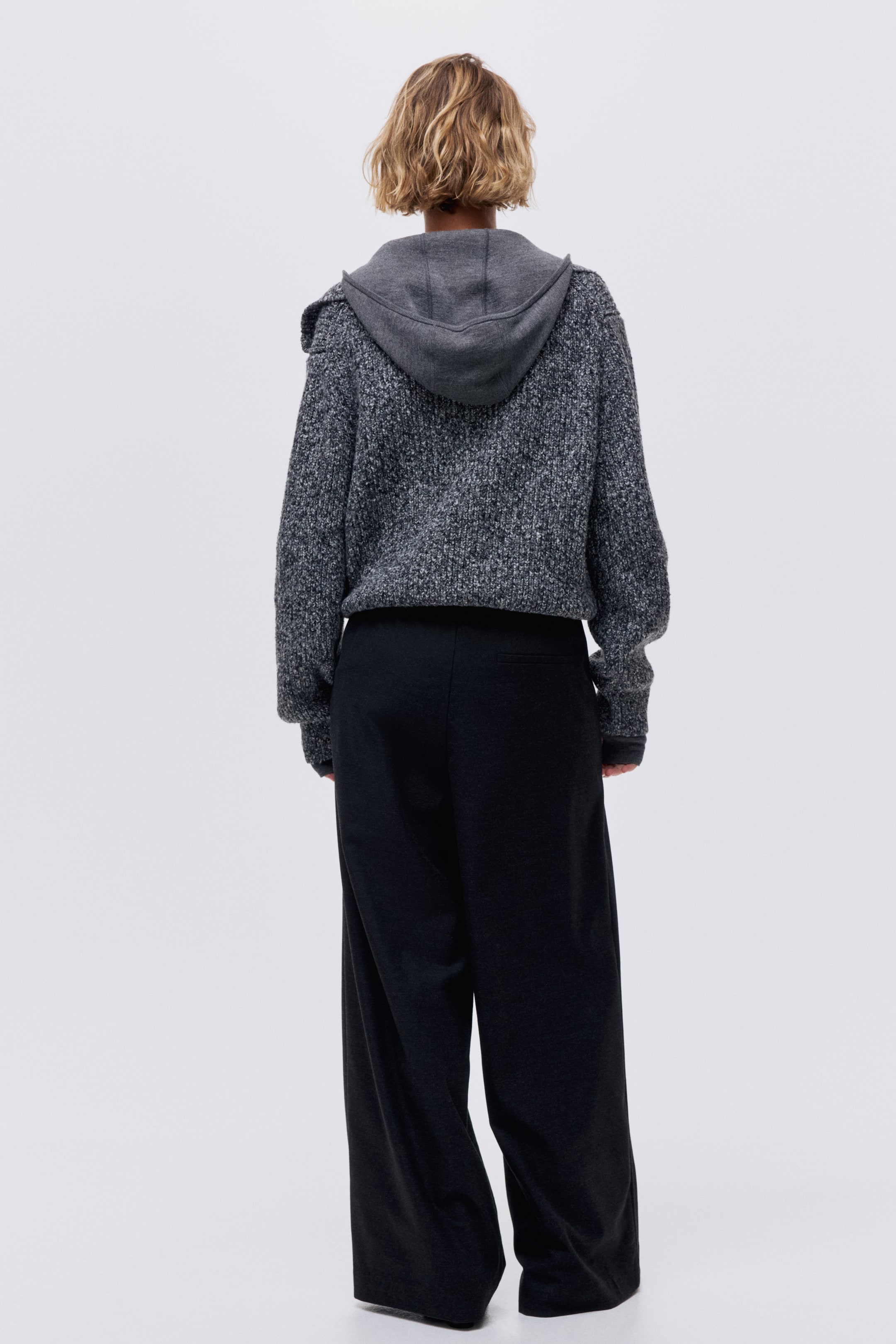 Näytä suurempi kuva: Pilkullinen harmaa neulottu hupullinen neuletakki, jossa on ribatut hihansuut, esitetään takaapäin. Se on kerrostettu tummanharmaan hupparin päälle ja yhdistetty mustiin leveälahkeisiin housuihin.