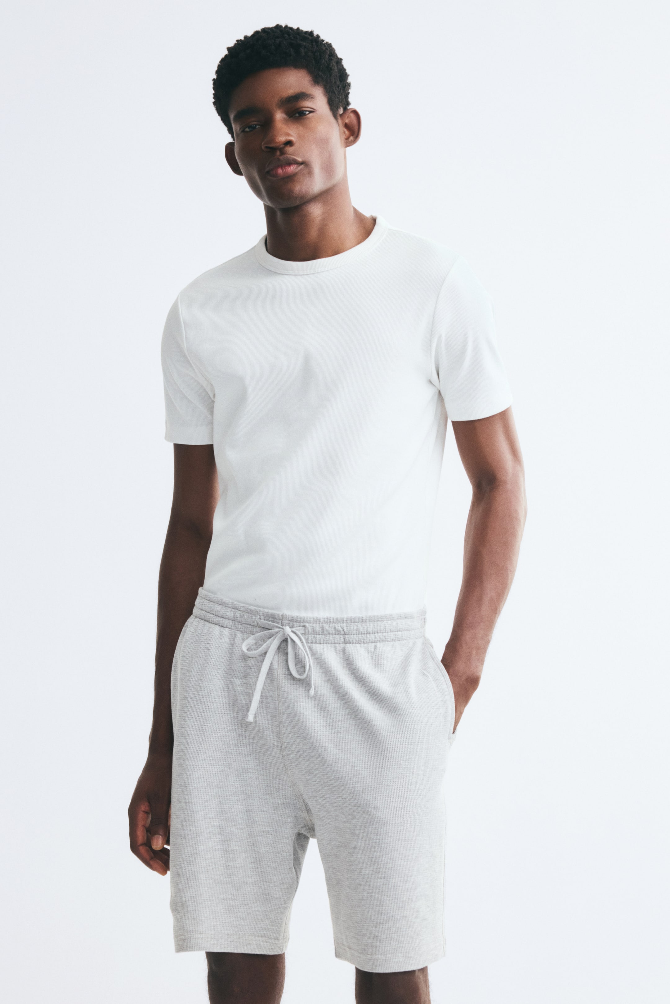View larger image: The model is wearing light grey waffled jersey shorts, featuring an elasticated waistband with a tied drawstring and a relaxed fit. The shorts are styled with a tucked-in white crewneck T-shirt, and the model is standing with one hand placed inside the welt side pocket.
