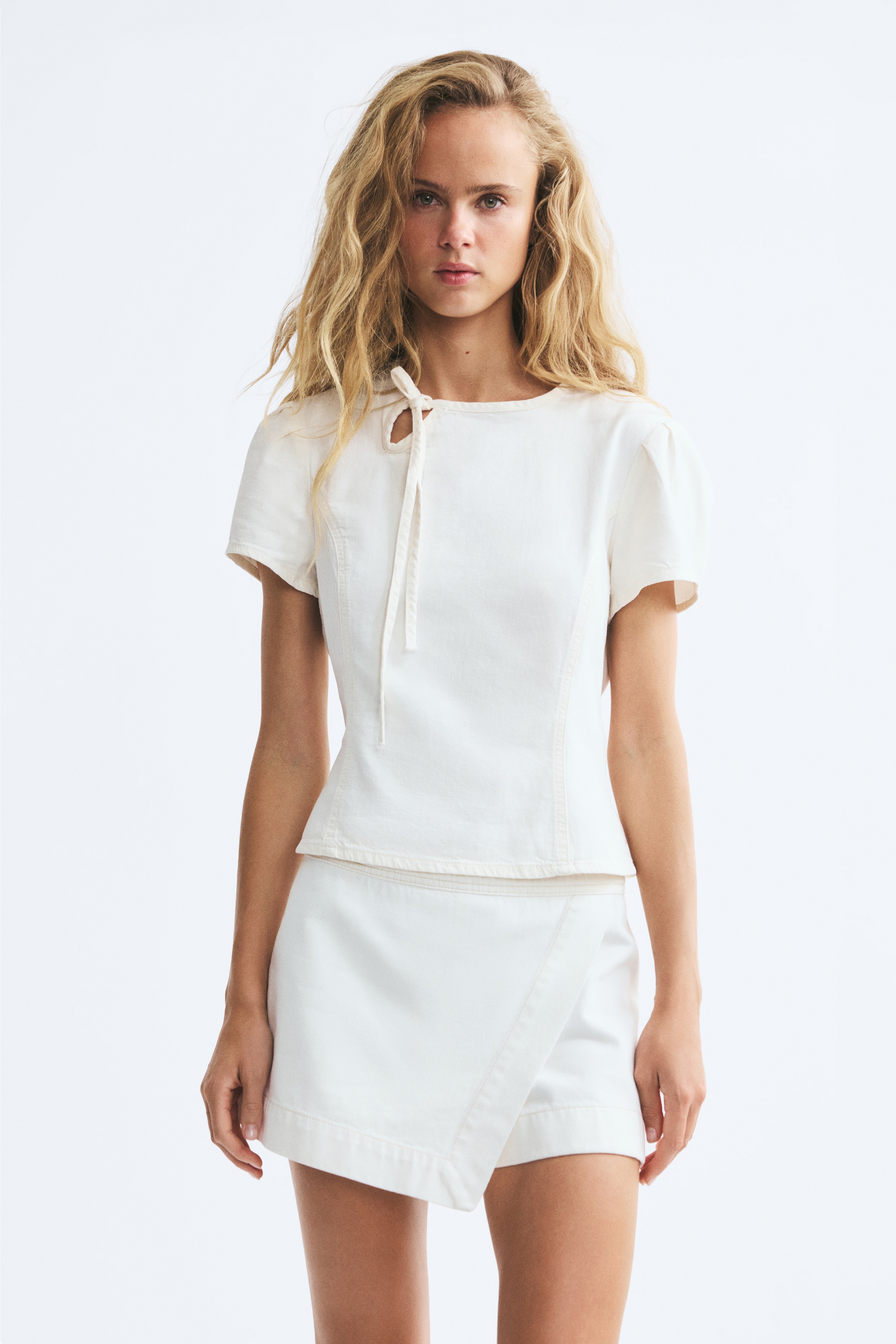 Má»t chiếc áo blouse trắng ôm dáng, kiá»u ngắn, ÄÆ°á»£c chụp từ phÃa trưá»c, vá»i cá» tròn có chi tiết khoét giá»t nưá»c nhá» và dây buá»c mảnh thắt nÆ¡ á» má»t bên. Ão có tay ngắn phá»ng và các ÄÆ°á»ng cắt may princess chạy dá»c phÃa trưá»c, tạo phom dáng rõ rà ng.