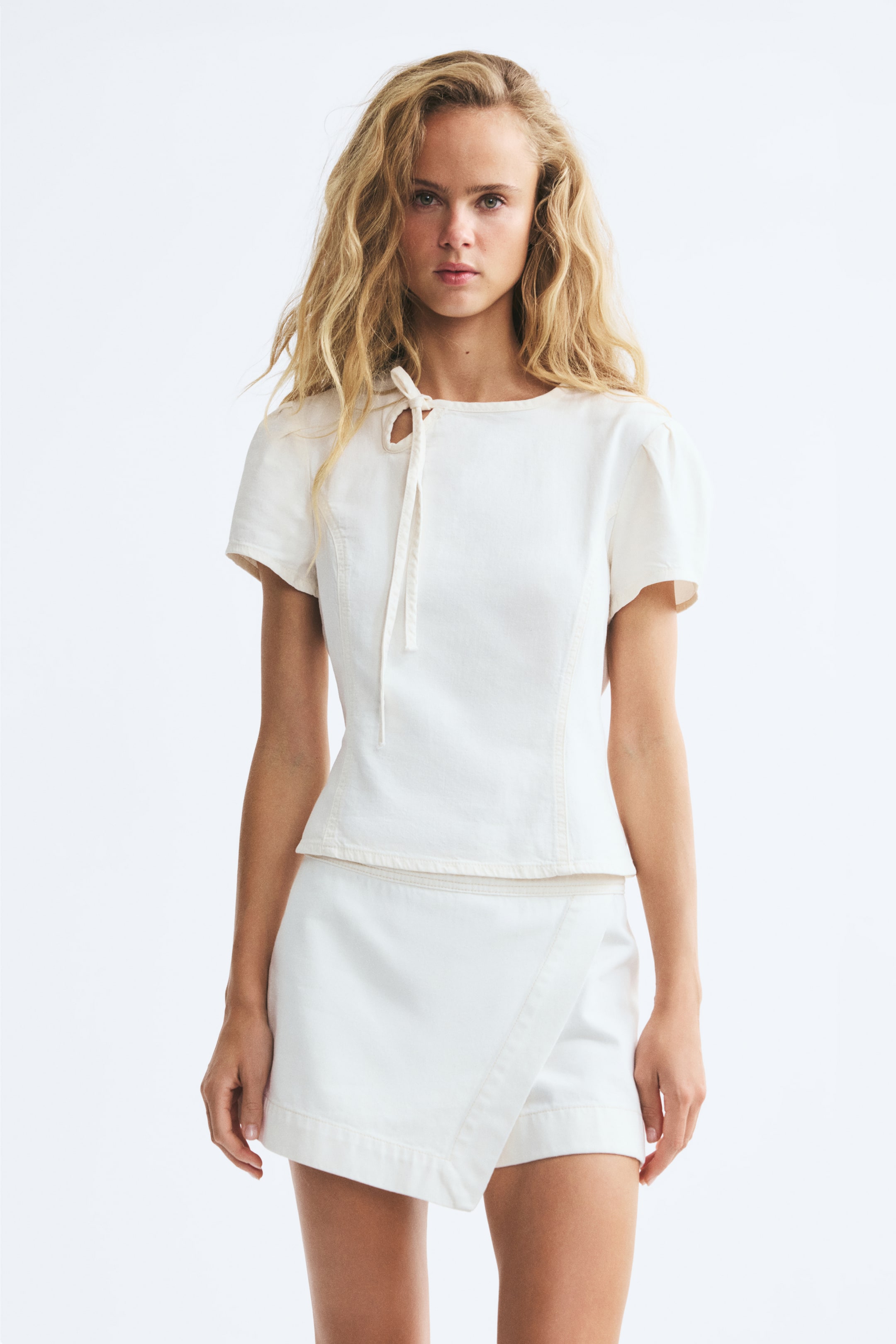 Xem ảnh lớn hơn: Người mẫu đang mặc một chiếc áo blouse ngắn, ôm dáng bằng cotton denim trắng, với cổ tròn và một lỗ khoét nhỏ kiểu keyhole cùng dây buộc mảnh ở một bên phía trước. Áo có tay ngắn phồng và các đường cắt princess chạy dọc thân trước, được phối cùng chân váy quần wrap skort denim trắng đồng bộ.