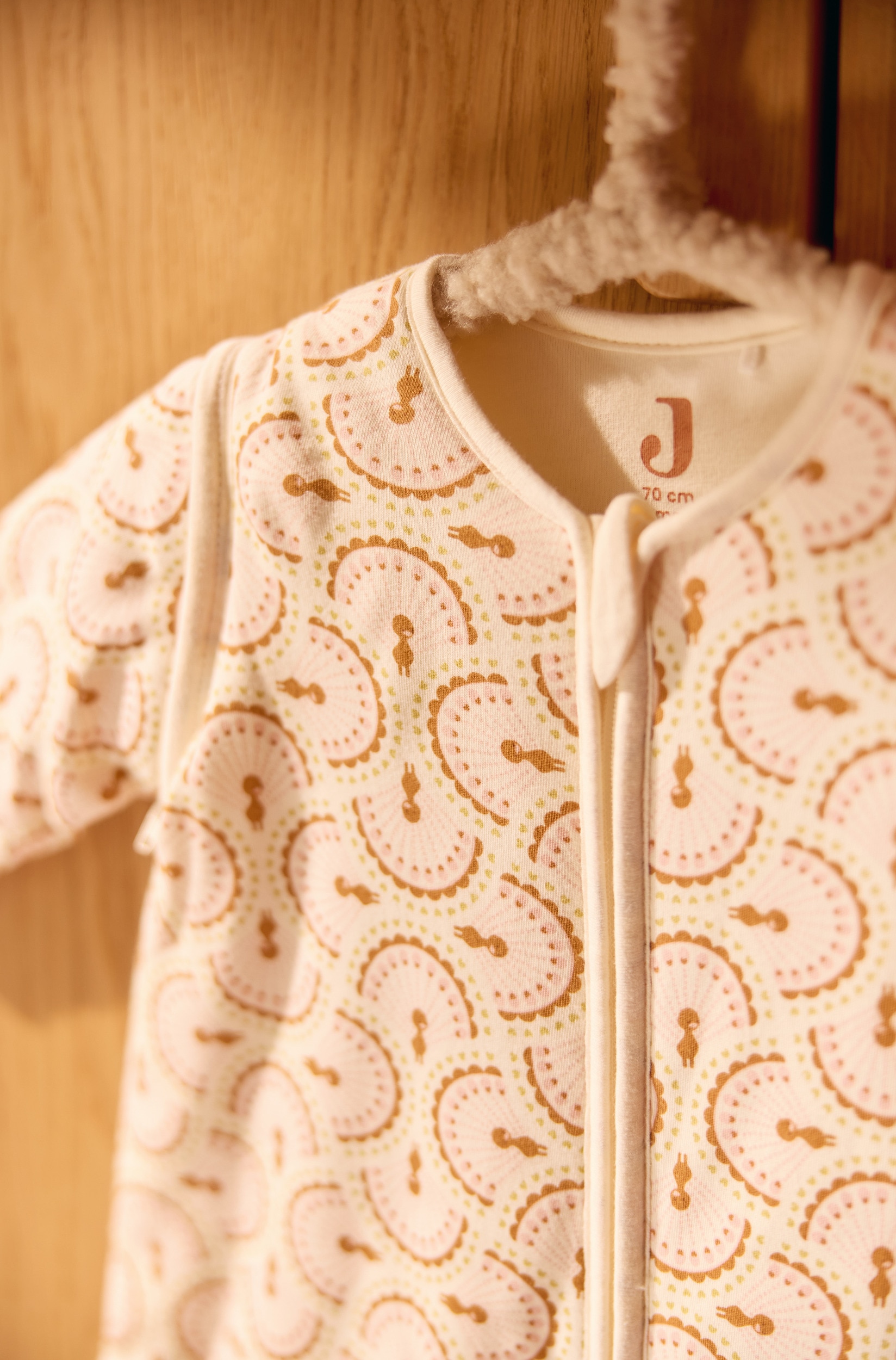 Une gigoteuse Ã  manches longues pour bÃ©bÃ© est posÃ©e Ã  plat, mettant en valeur un motif rÃ©pÃ©tÃ© sur toute la surface de motifs en Ã©ventail semi-circulaires beige sur un fond blanc cassÃ©. La gigoteuse prÃ©sente une fermeture Ã©clair intÃ©grale centrale sur le devant, un col rond et de longues manches longues.