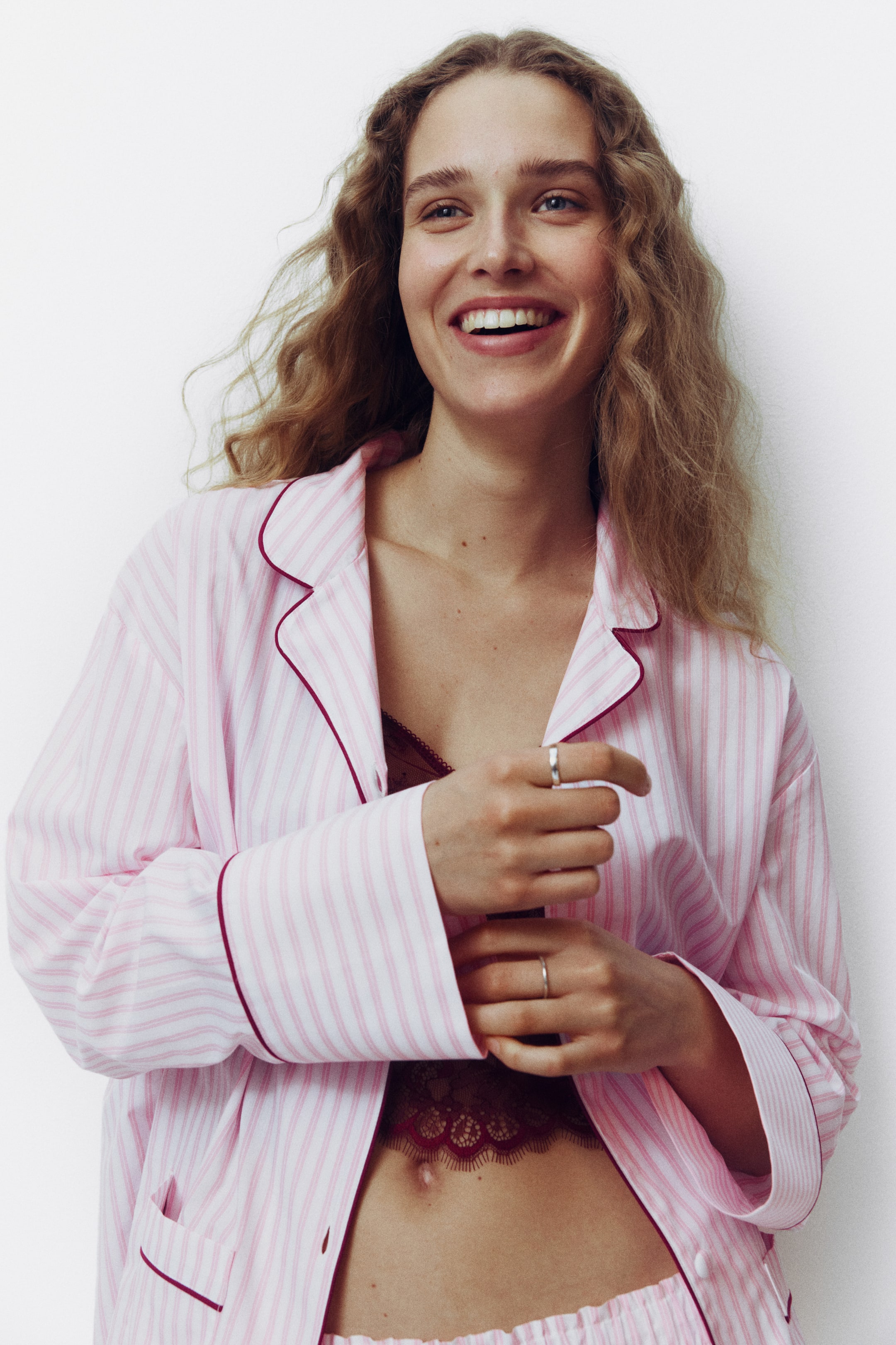 View larger image: Presenting a pink and white striped pajama set, which includes a collared top with notch lapels and red piping detailing along the collar, lapels, front opening, and cuffs. The top is styled open, showcasing a burgundy lace bralette, while the matching striped pajama shorts are visible at the waist.