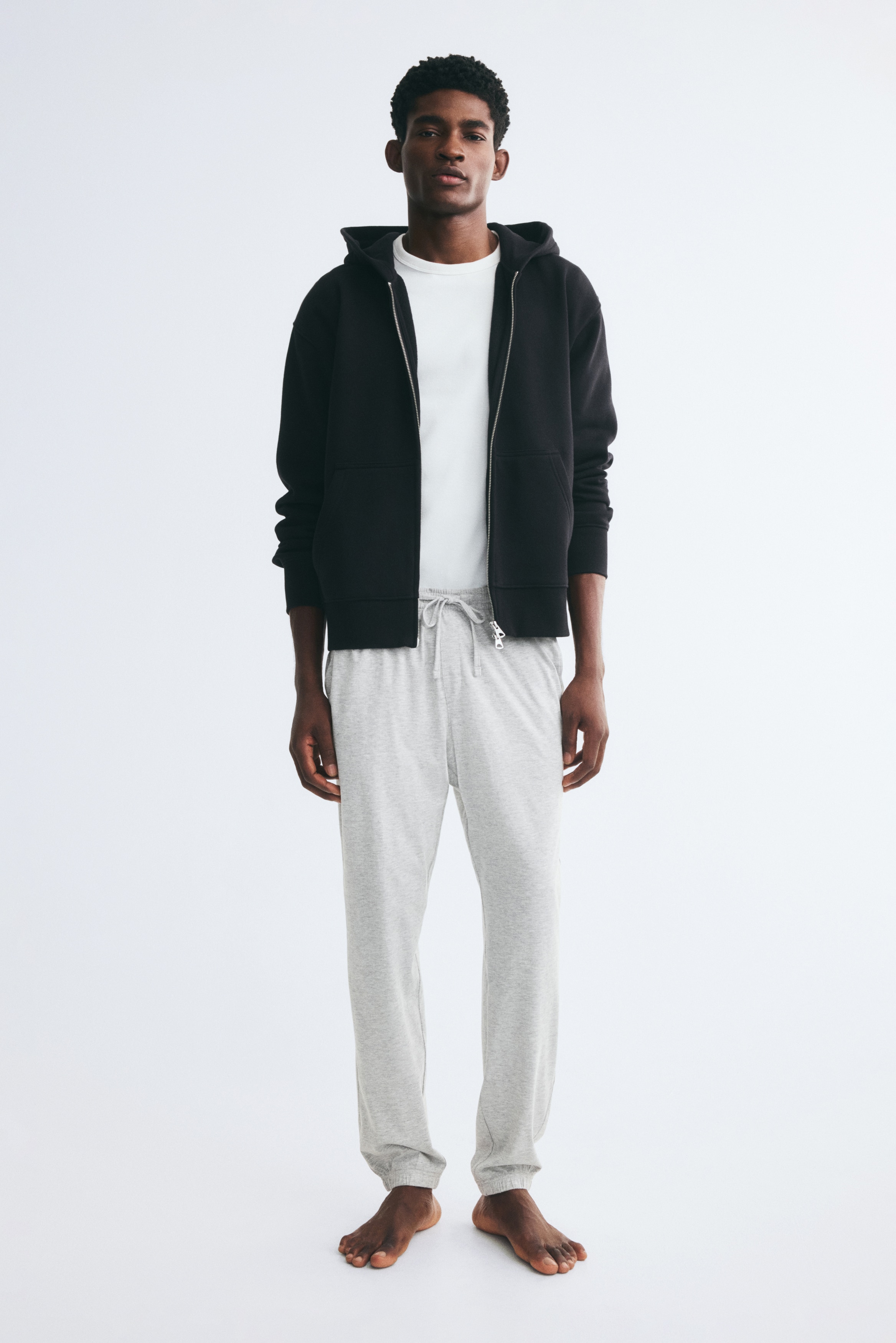Deux paires de pantalons de survêtement en jersey de coton sont présentées face à l'AVANT. La paire gris clair, positionnée devant, présente une taille élastiquée avec cordon de serrage, un détail de braguette cousue, des poches latérales et des ourlets côtelés. Une paire bleu foncé est partiellement visible derrière, affichant également des poches latérales et des ourlets côtelés.