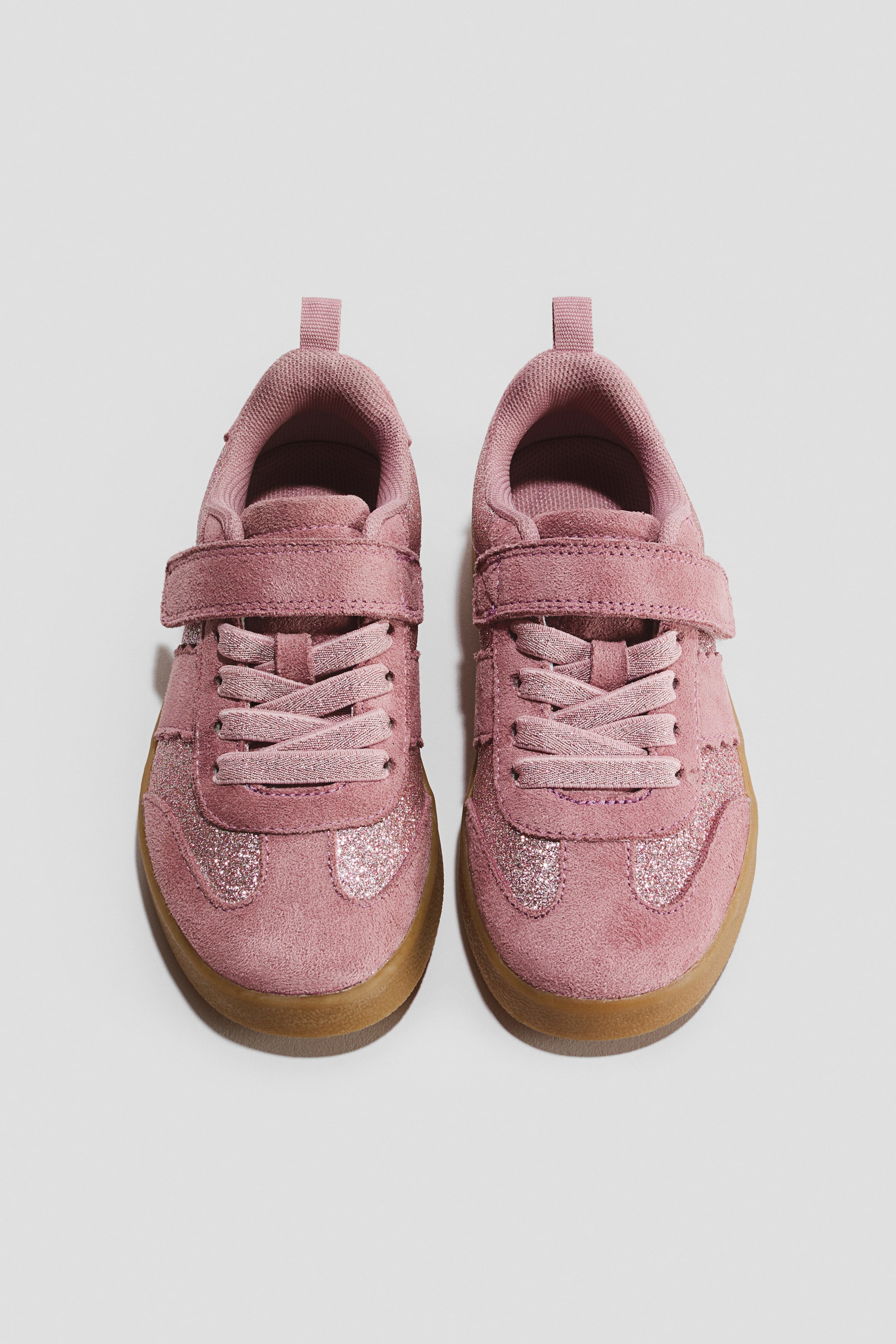 View larger image: A pair of retro-style sneakers is shown from a top-down perspective, featuring a dusty pink suede upper with glittery panels on the toe box. The sneakers feature a single hook-and-loop strap over elasticated laces, a padded collar with a heel pull tab, and a contrasting brown gum sole.