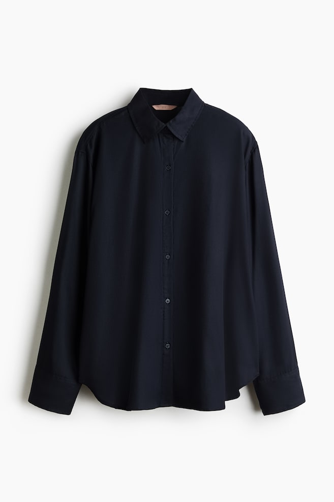 A loose-fit, dark navy blue shirt is hanging in a front-facing view, featuring a collar, a button-down front, and gently dropped shoulders. The shirt is displaying long sleeves with wide button cuffs and a curved, rounded hem. - 1