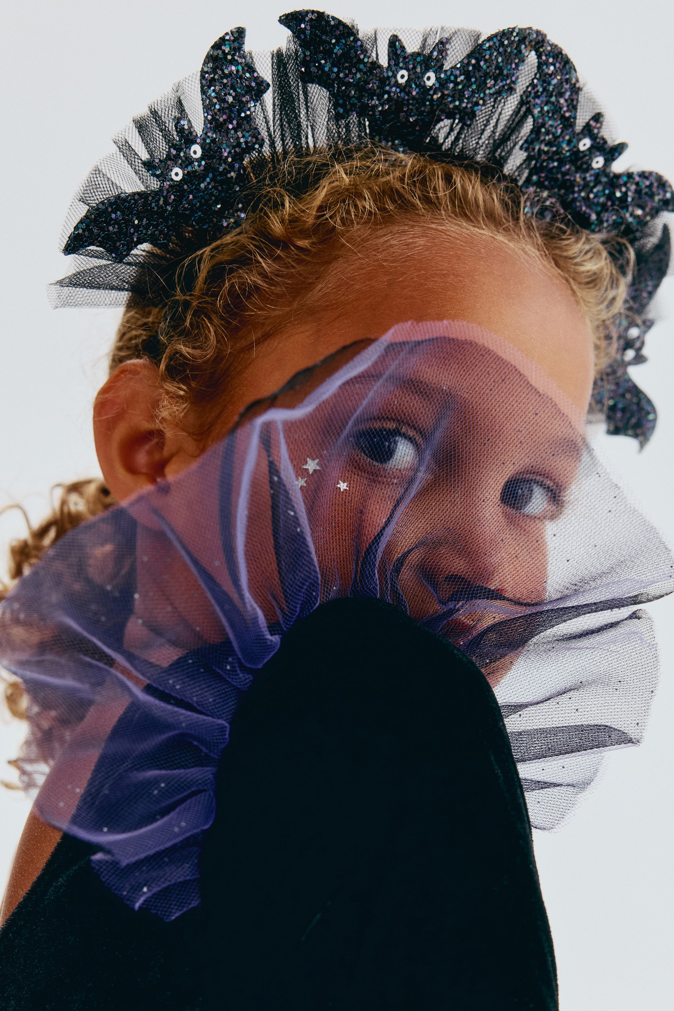Grotere afbeelding bekijken: Het product toont een zwarte glittervleermuizenkroon met vleermuizen met witte ogen, bevestigd op een gerimpelde zwarte tulebasis. Een doorschijnende paarse mesh-ruches, versierd met zilveren sterren, bedekt gedeeltelijk het onderste deel van het gezicht.