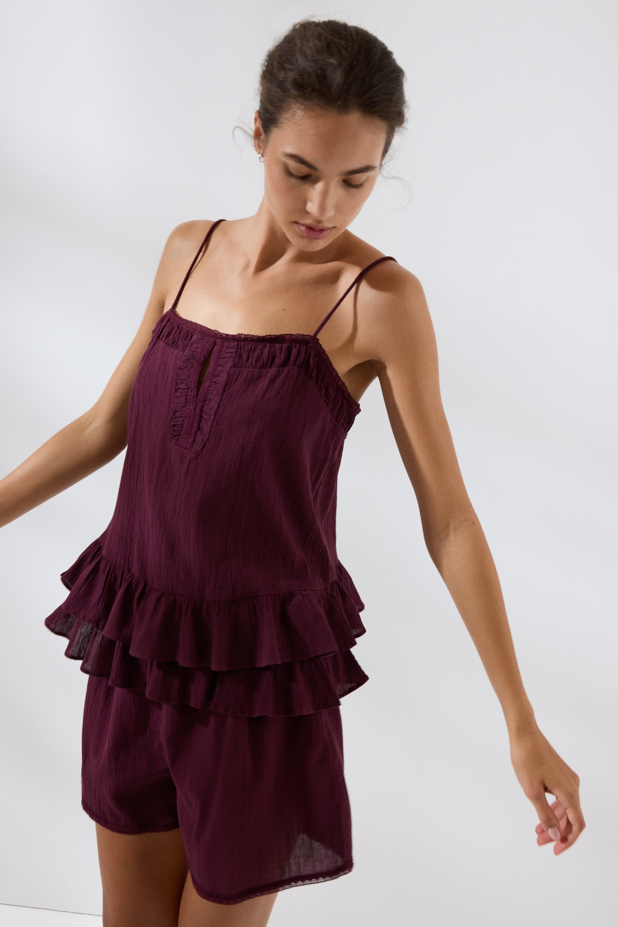 Größeres Bild ansehen: Der Pyjama aus weinroter, geknitterter Baumwolle, bestehend aus einem Oberteil und Shorts, wird von dem Modell getragen. Das Oberteil zeichnet sich durch einen geraden Ausschnitt, Spitzenbesatz, vordere Raffungen, einen Schlüssellochausschnitt, schmale Träger und einen doppelten Rüschenbesatz am Saum aus. Die Shorts verfügen über einen Spitzenbesatz an den Säumen.