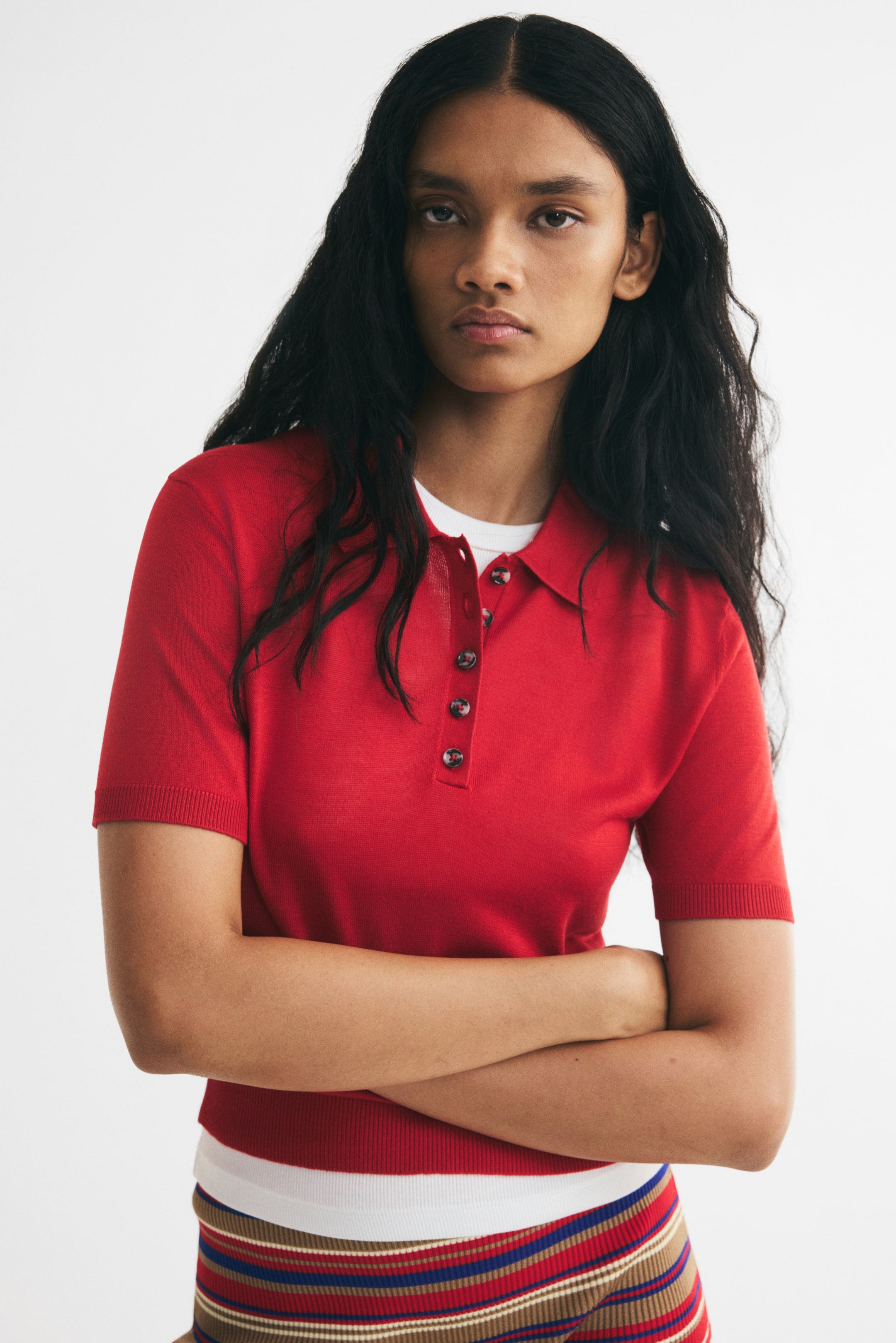 Ampliar la imagen: Una playera polo tejida roja con cuello, una tapeta parcialmente desabotonada y mangas cortas con puños acanalados es llevada por la modelo con los brazos cruzados. La playera polo está combinada sobre una playera blanca de cuello redondo y acompañada de una falda tejida a rayas.