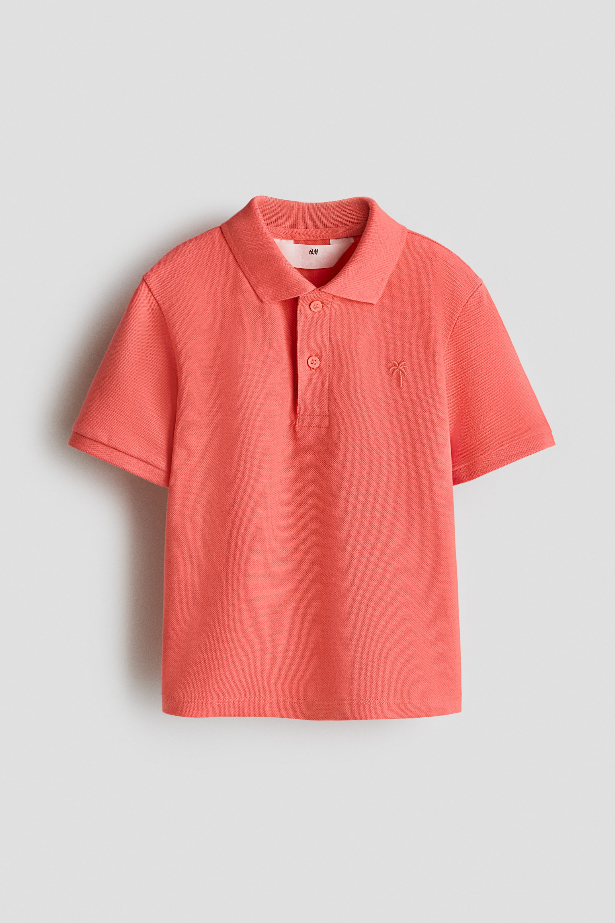 View larger image: A coral short-sleeved polo shirt is laid flat, featuring a classic collar, a two-button placket with matching buttons and an embroidered palm tree on the left chest.