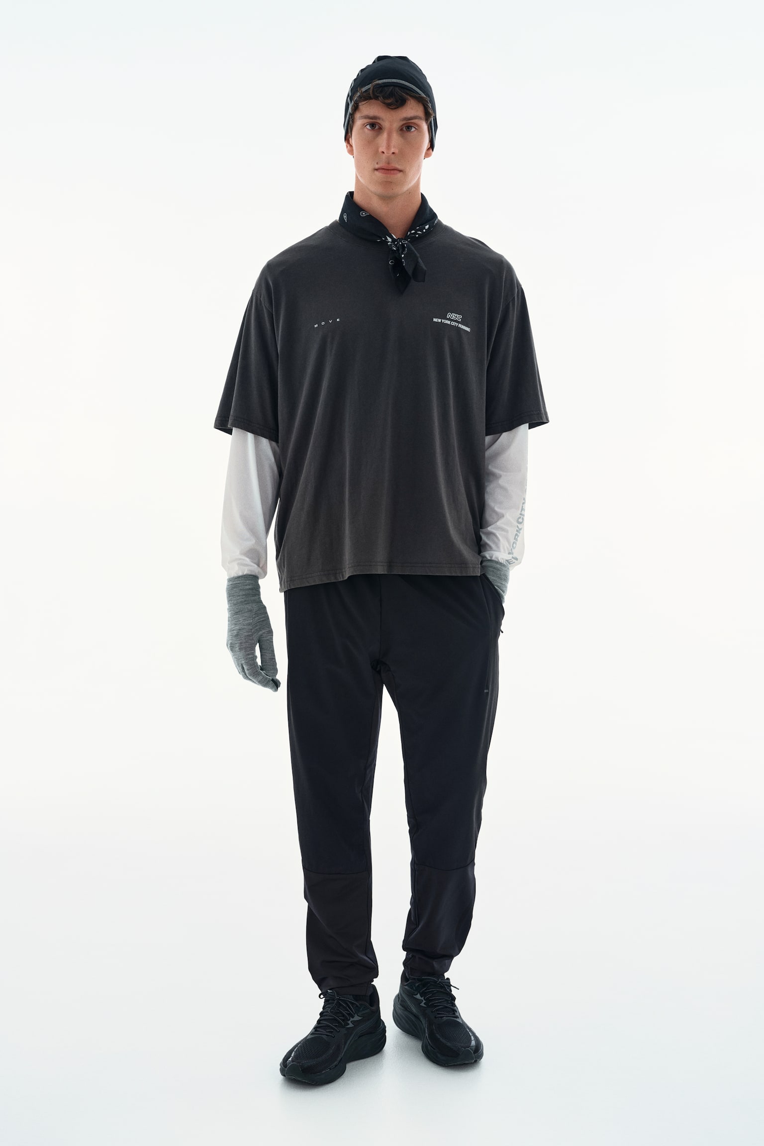 A dark grey oversized short-sleeved T-shirt with small white text graphics on the chest is worn by the model. The T-shirt is layered over a white long-sleeved top and styled with black athletic trousers, a black beanie, a black neck bandana, and grey gloves. - 1