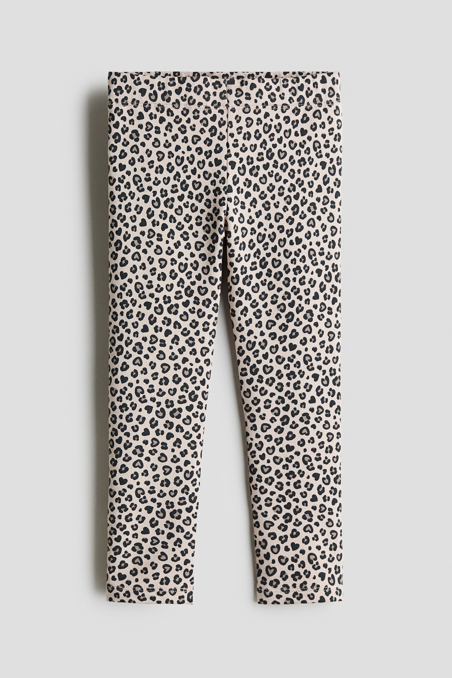 Leggings aus hellbeigem Baumwoll-Jersey mit einem Allover-Leopardenmuster in Schwarz und Grau, das aus kleinen Herzformen besteht. Die Vorderansicht zeigt den flachen, elastischen Bund und die schmal geschnittenen Beine.