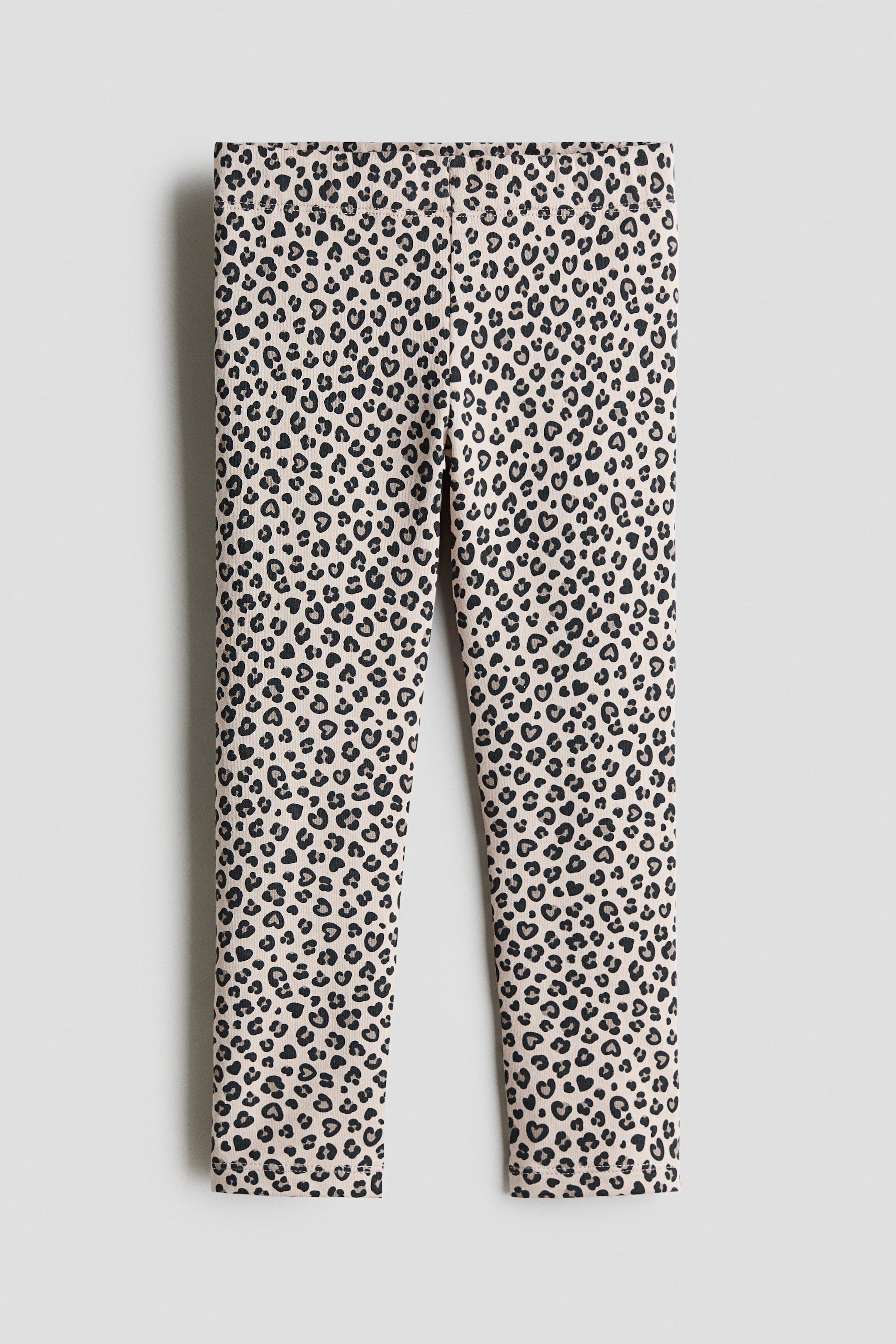 Größeres Bild anzeigen: Leggings aus hellbeigem Baumwolljersey mit einem allover Leopardenmuster in Schwarz und Grau, das aus kleinen Herzformen besteht. Die Vorderansicht zeigt den flachen, elastischen Bund und die schmal geschnittenen Beine.