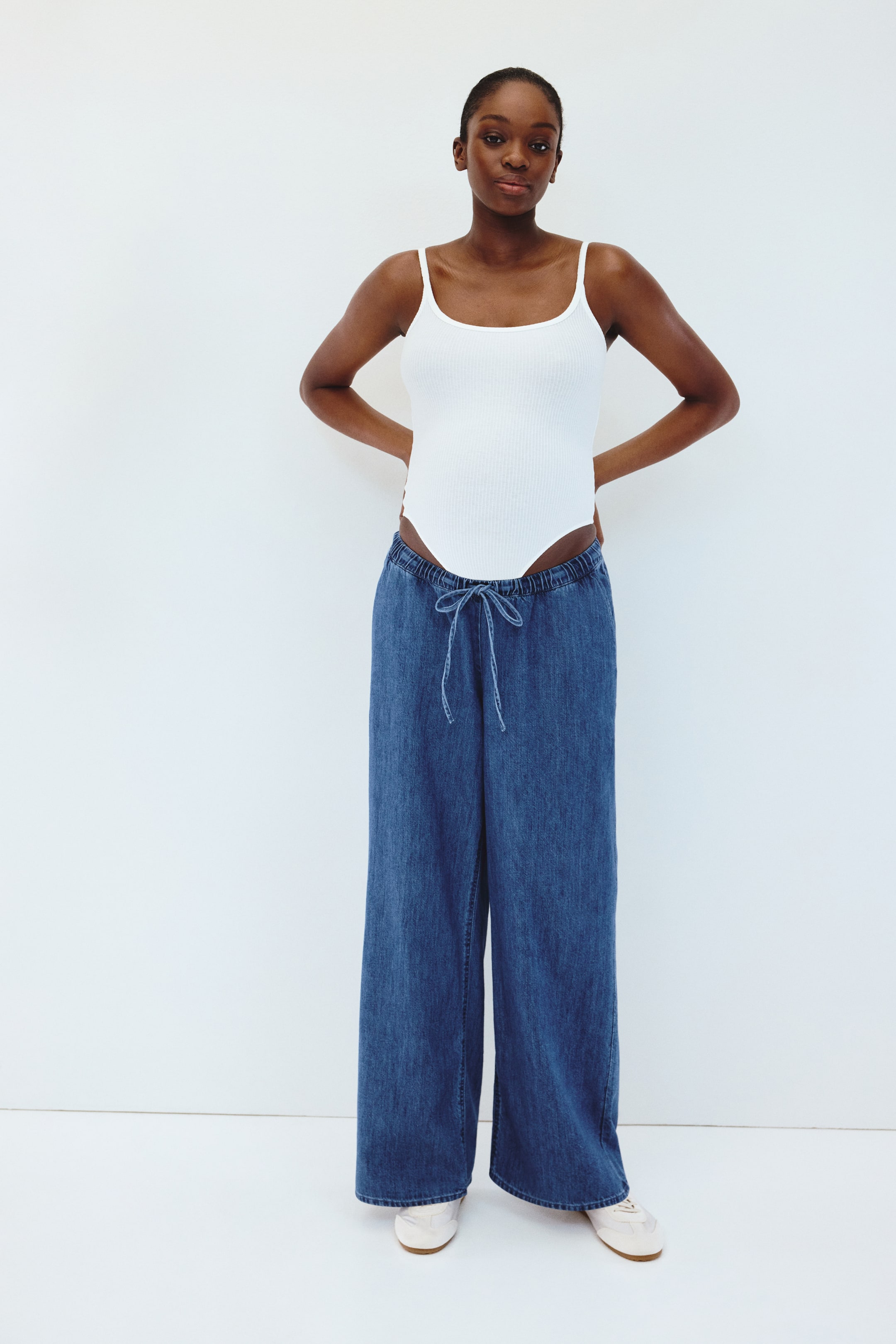 View larger image: A white ribbed scoop-neck bodysuit with spaghetti straps is worn, shown from the front, tucked into blue wide-leg trousers with a drawstring waist and a relaxed fit.