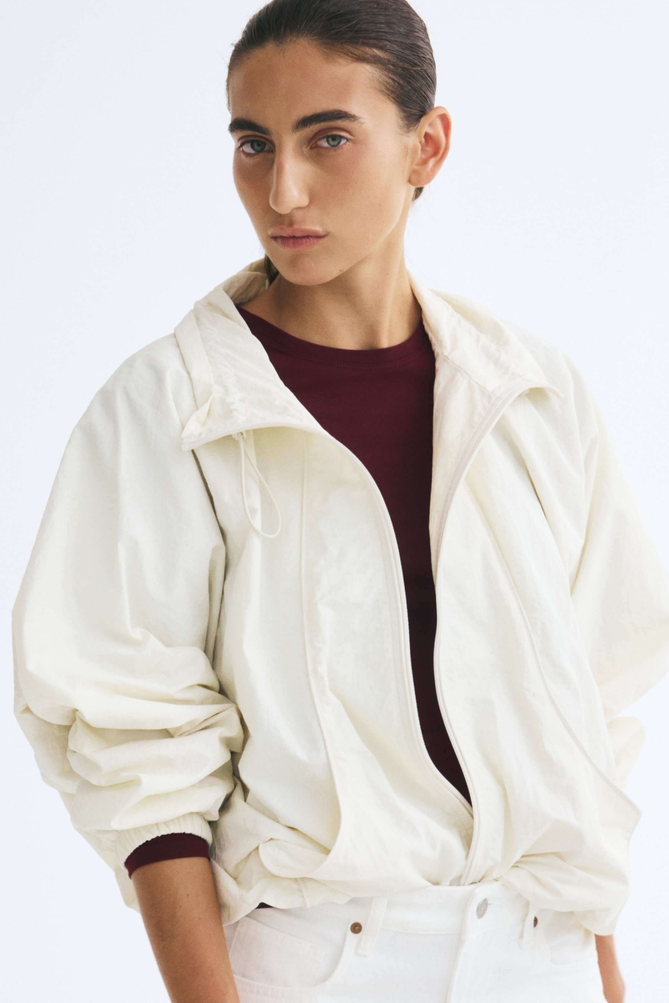 View larger image: The model is wearing a fitted burgundy top with a round neckline and long sleeves. The top is styled as a base layer under an open, off-white lightweight jacket.