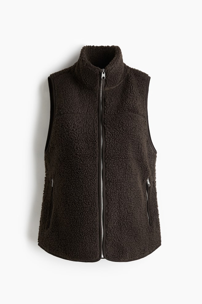 A dark brown gilet is presented from the FRONT, featuring a textured, soft teddy material. It is designed with a stand collar, a full-length silver zipper down the center, and two vertical zipped pockets on the lower front. - 1