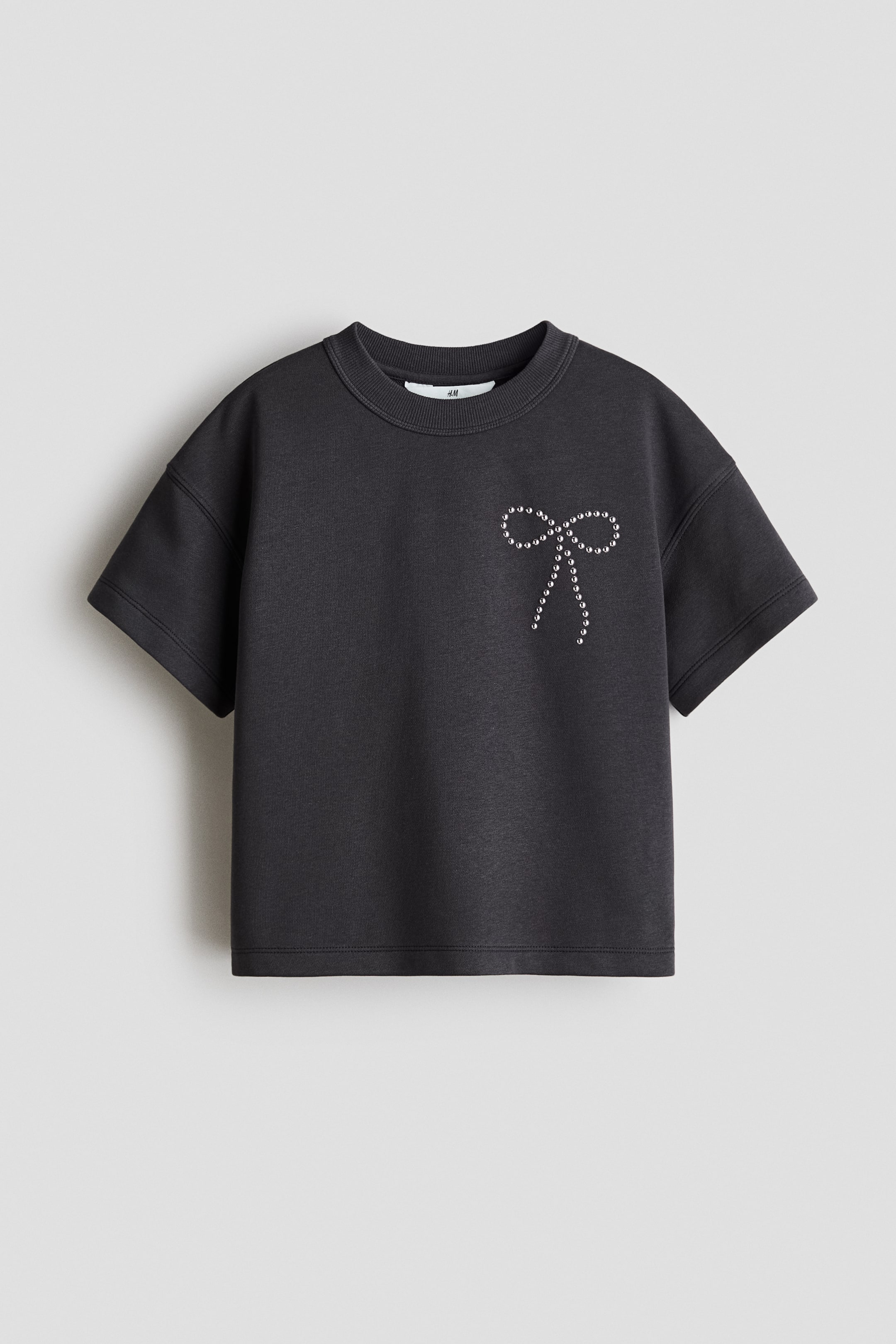 Voir une image plus grande: Un T‑shirt en jersey gris foncé est présenté de face, affichant une coupe ample, une encolure ronde à bordure côtelée et des épaules légèrement tombantes. Un motif en forme de nœud, composé de petits clous brillants, orne le côté gauche de la poitrine.