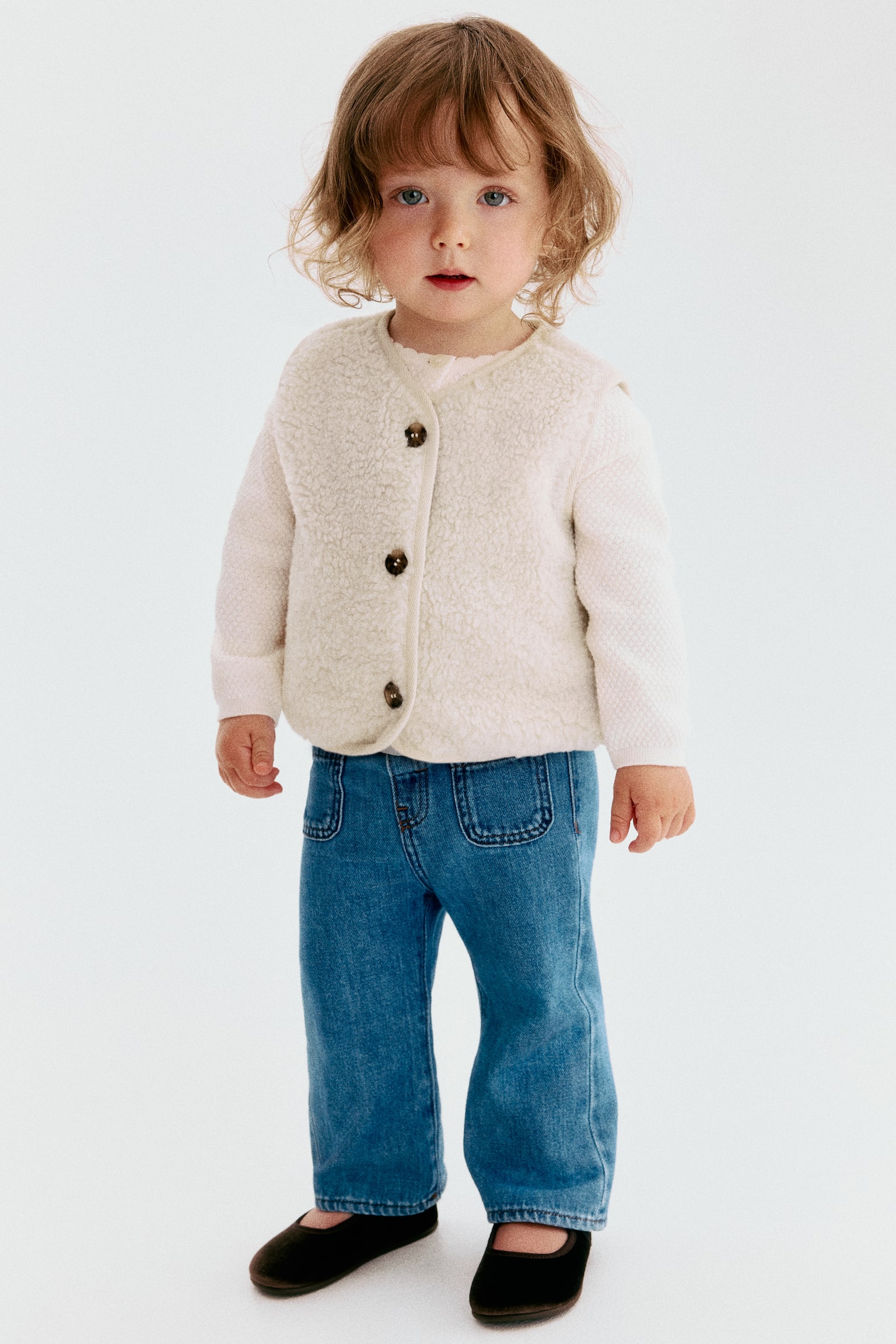 A gilet in soft cream teddy is shown from the front, featuring a round neckline and dark brown buttons down the front. Twill binding details the edges and placket, with a white long-sleeved top worn underneath. - 1