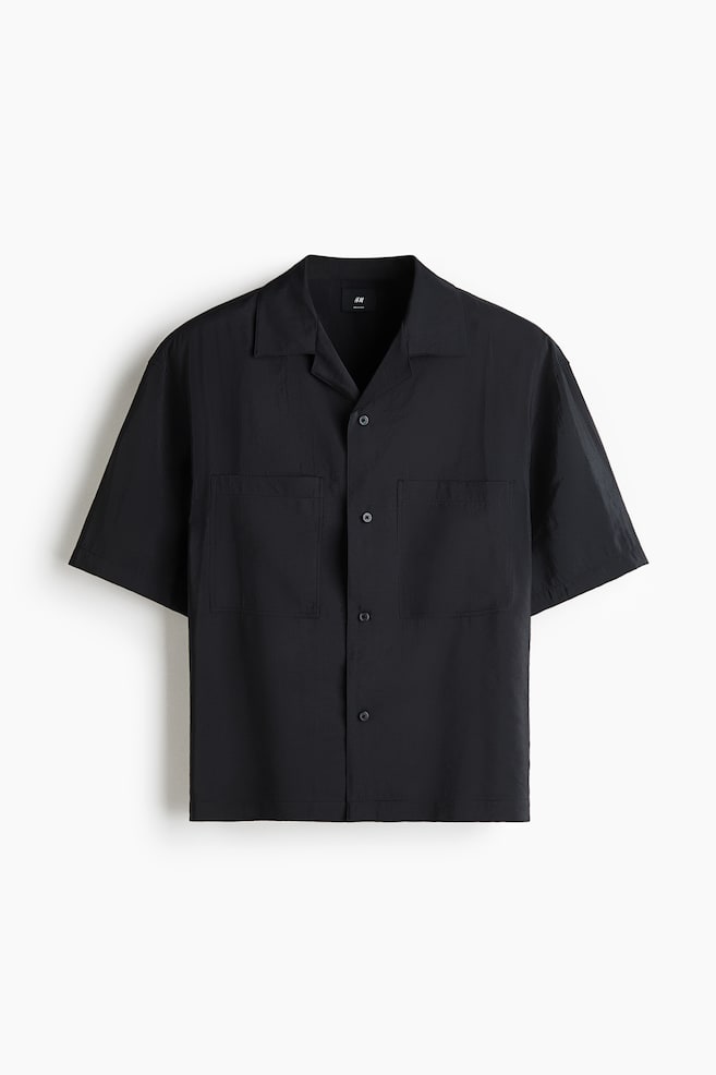A black short-sleeved shirt featuring a camp collar, a button-front closure with black buttons, and two patch pockets on the chest. The shirt showcases a slightly textured fabric. - 1