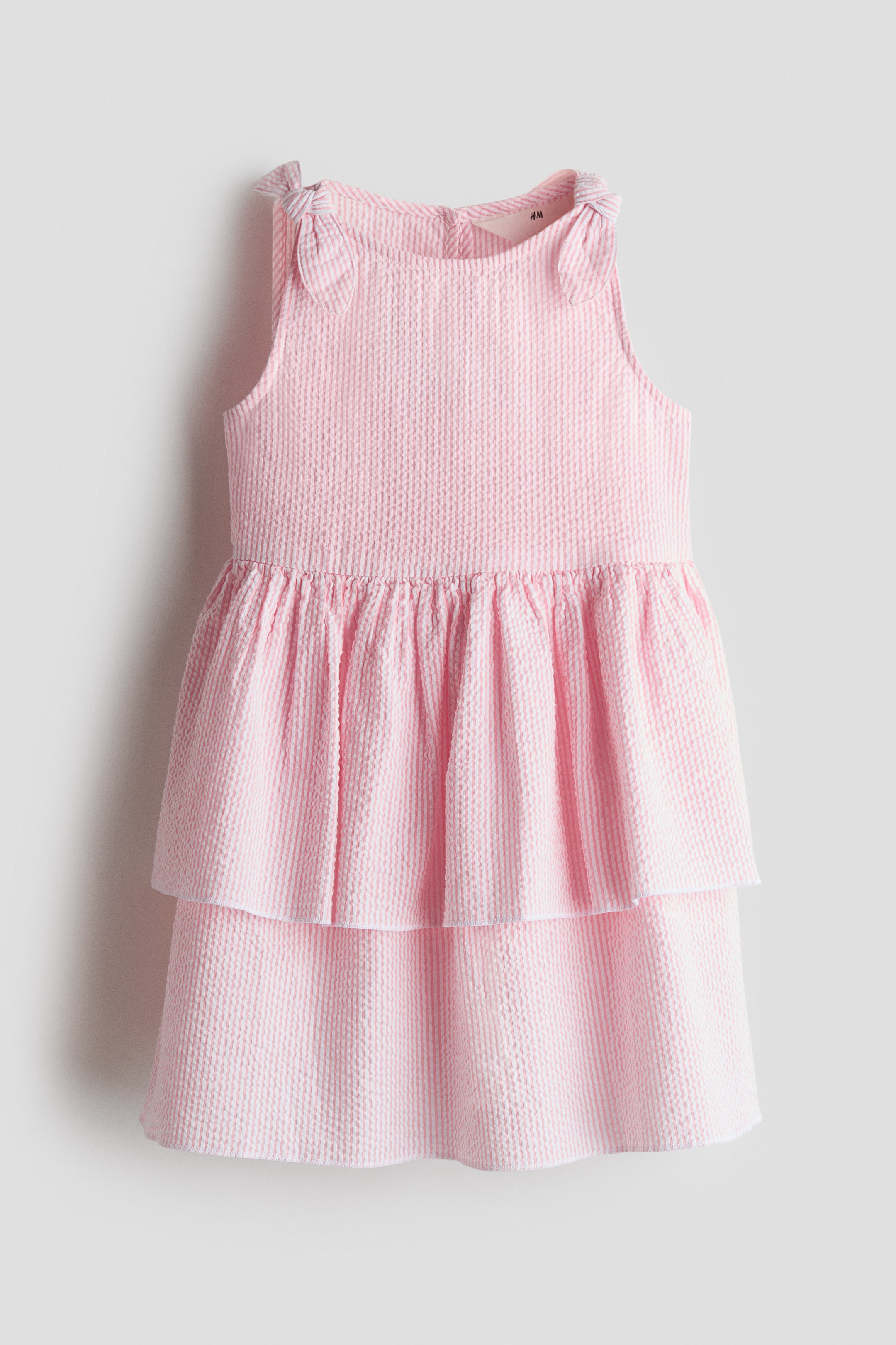 View larger image: A light pink and white striped seersucker dress is showcasing a tiered skirt design. It features shoulder straps tied into bows and a round neckline.