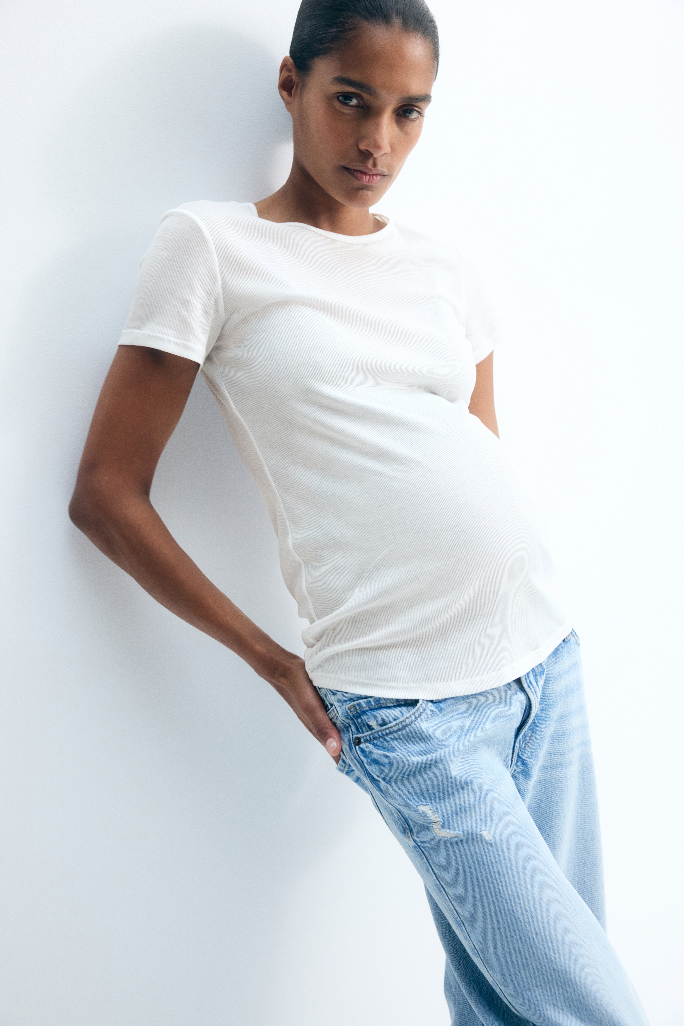 A white maternity T-shirt is shown from the front, featuring a fine ribbed texture, short sleeves, and a round neckline. The top showcases a fitted silhouette with a curved hemline designed to accommodate a pregnancy bump.