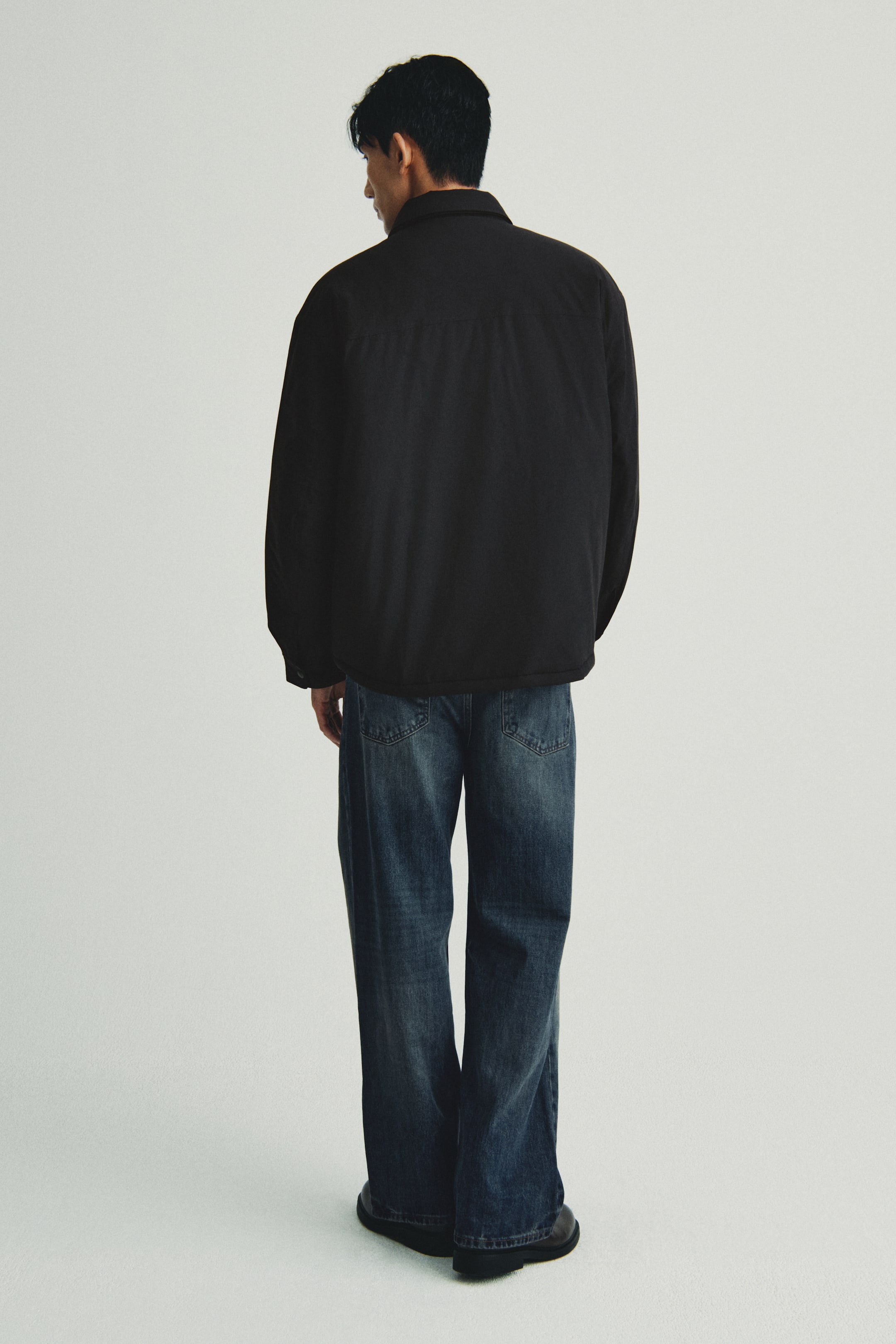 이미지 확대 보기: 검은색의 헐렁한 재킷이 뒤에서부터 소개되고 있으며, 칼라와 상부 뒤쪽을 가로지르는 요크가 특징입니다. 어두운 청색의 와이드 레그 청바지와 스타일링되어 있으며, 이 역시 뒤에서 보여지며, 눈에 띄는 뒷주머니가 있습니다.