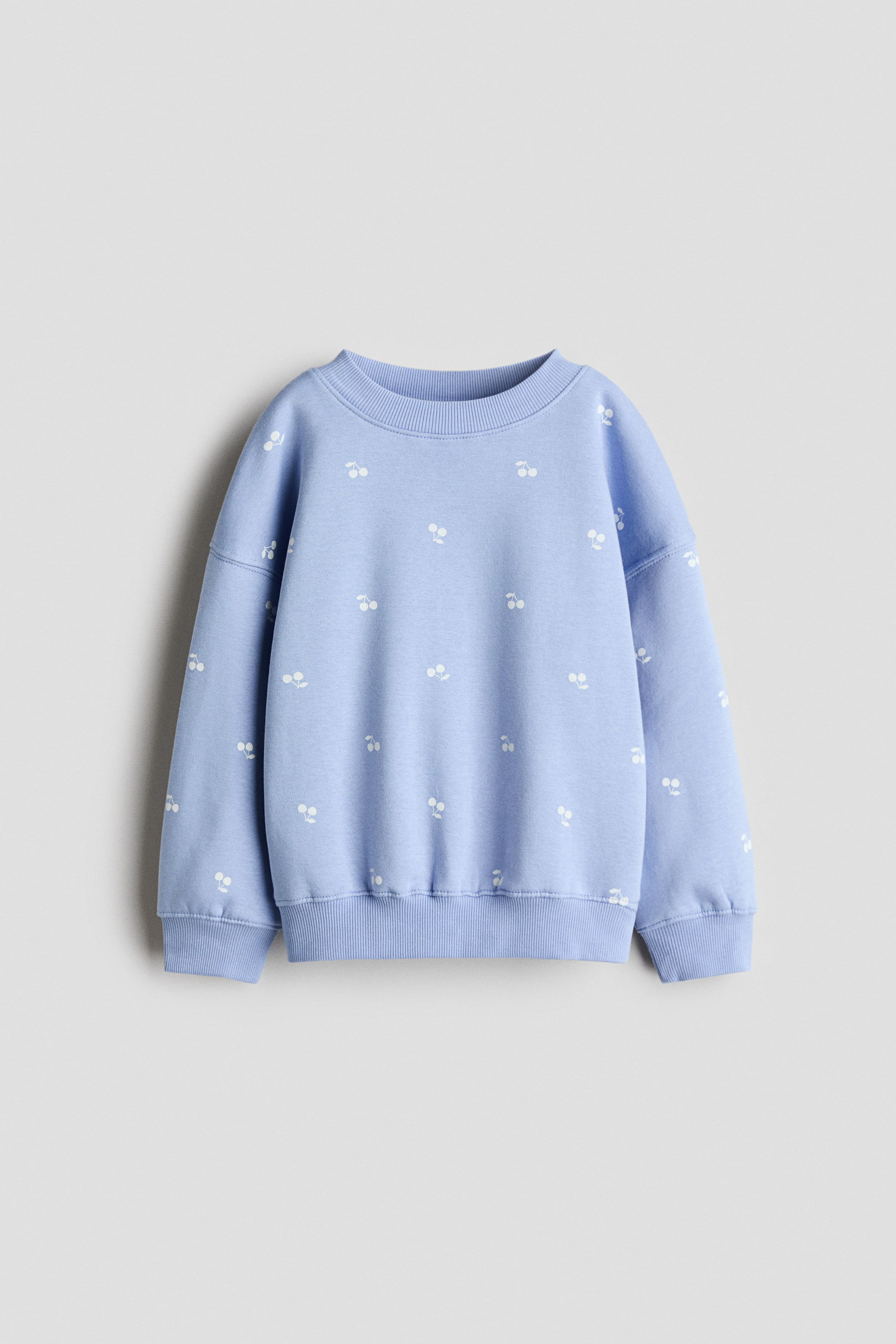 View larger image: A light blue sweatshirt is shown from the front, featuring a round, rib-trimmed neckline, dropped shoulders, and long sleeves. The sweatshirt is patterned with small white cherry motifs and finished with ribbing at the cuffs and hem.