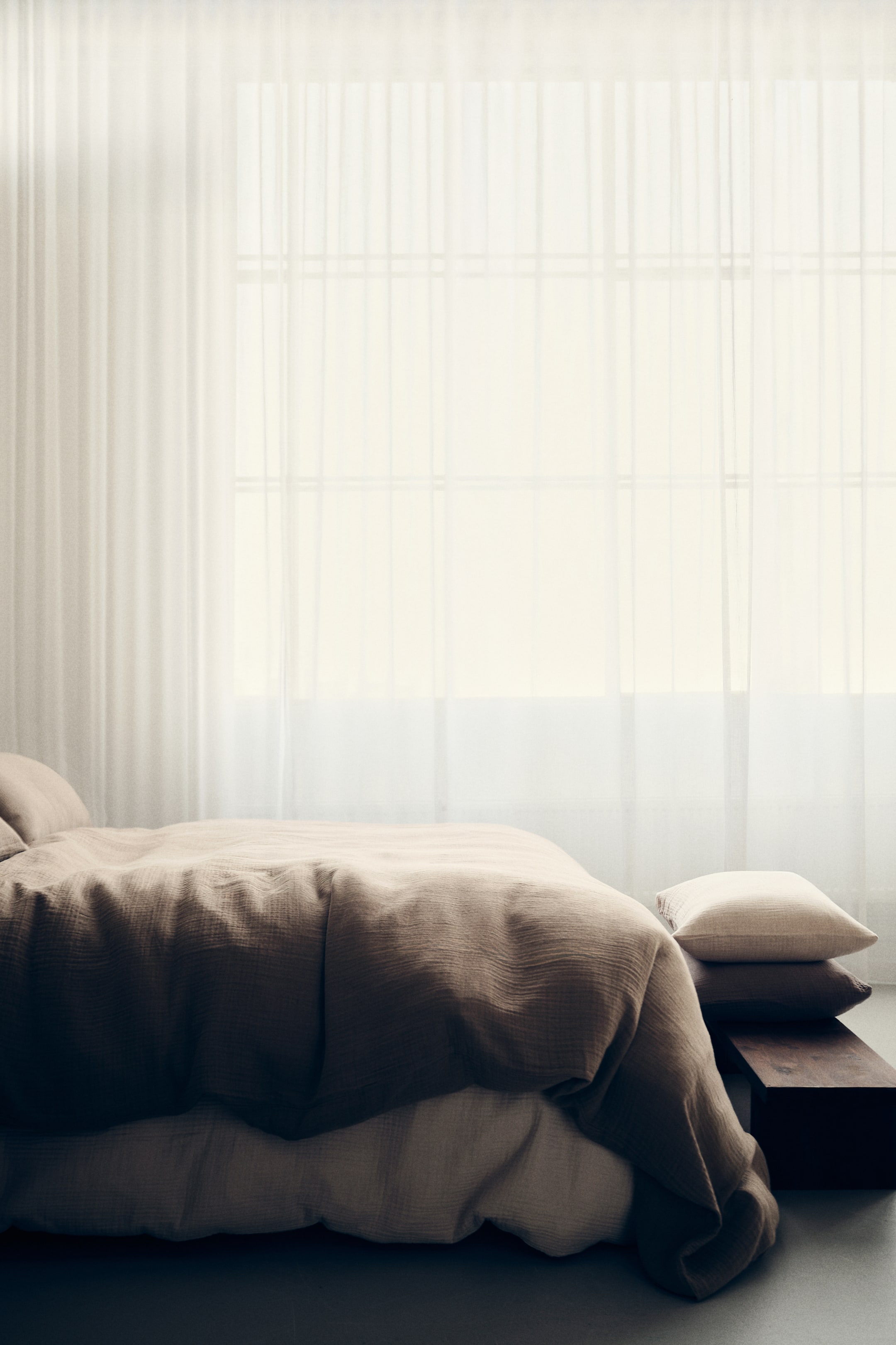 Ingrandisci l'immagine: Un set di biancheria da letto con una texture increspata di colore marrone chiaro comprende un copripiumino e cuscini coordinati. Sotto il piumino è visibile uno strato di lenzuola di colore più chiaro, e due cuscini di colore più chiaro sono impilati ai piedi del letto, appoggiati su un blocco di legno scuro.