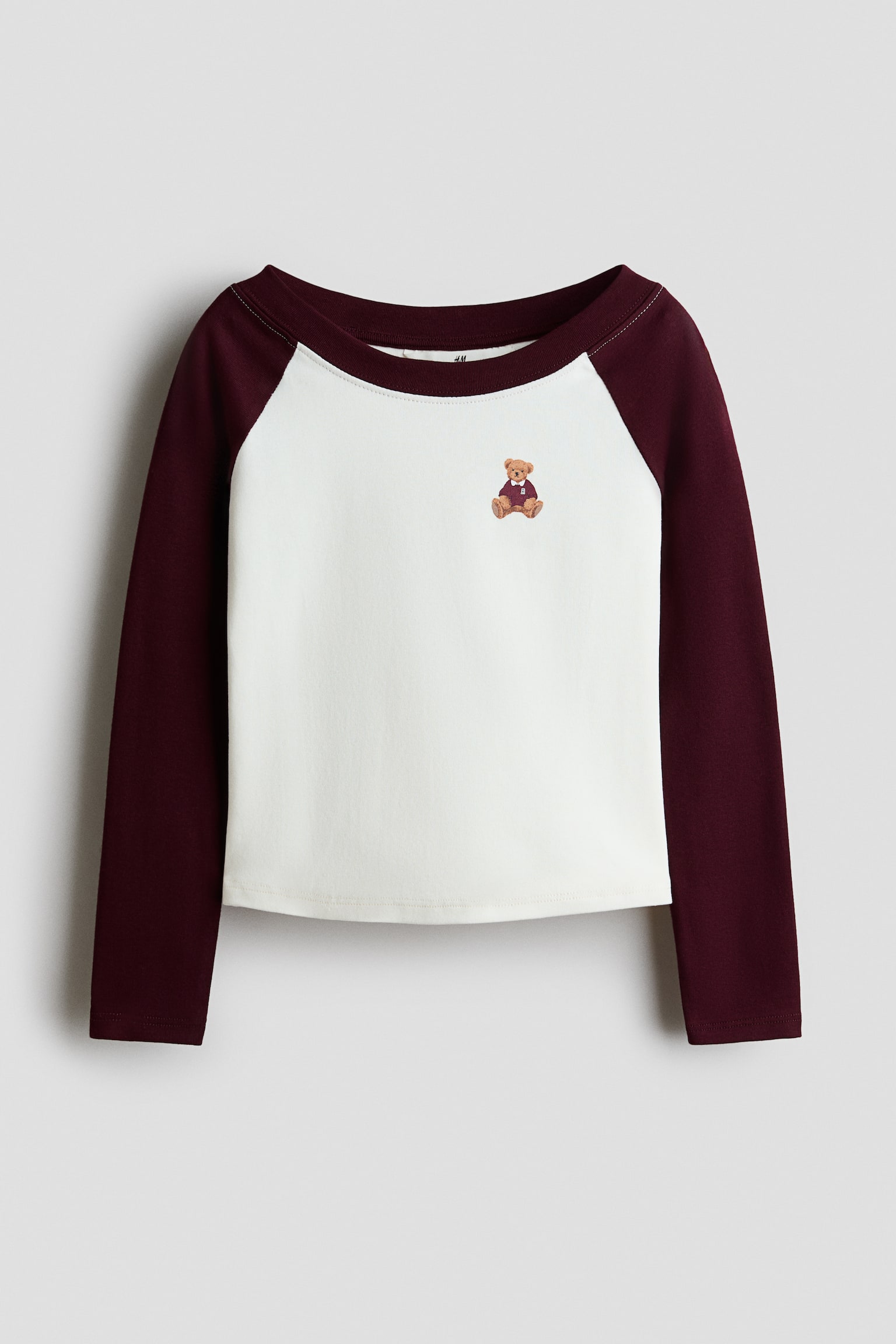 Een T-shirt met lange raglanmouwen wordt van voren getoond, met een crÃ¨mekleurig lichaam en een kleine teddybeerafbeelding op de linkerborst bovenaan. Het T-shirt heeft contrasterende lange raglanmouwen in donker bordeaux en een bijpassende ronde halsafwerking.
