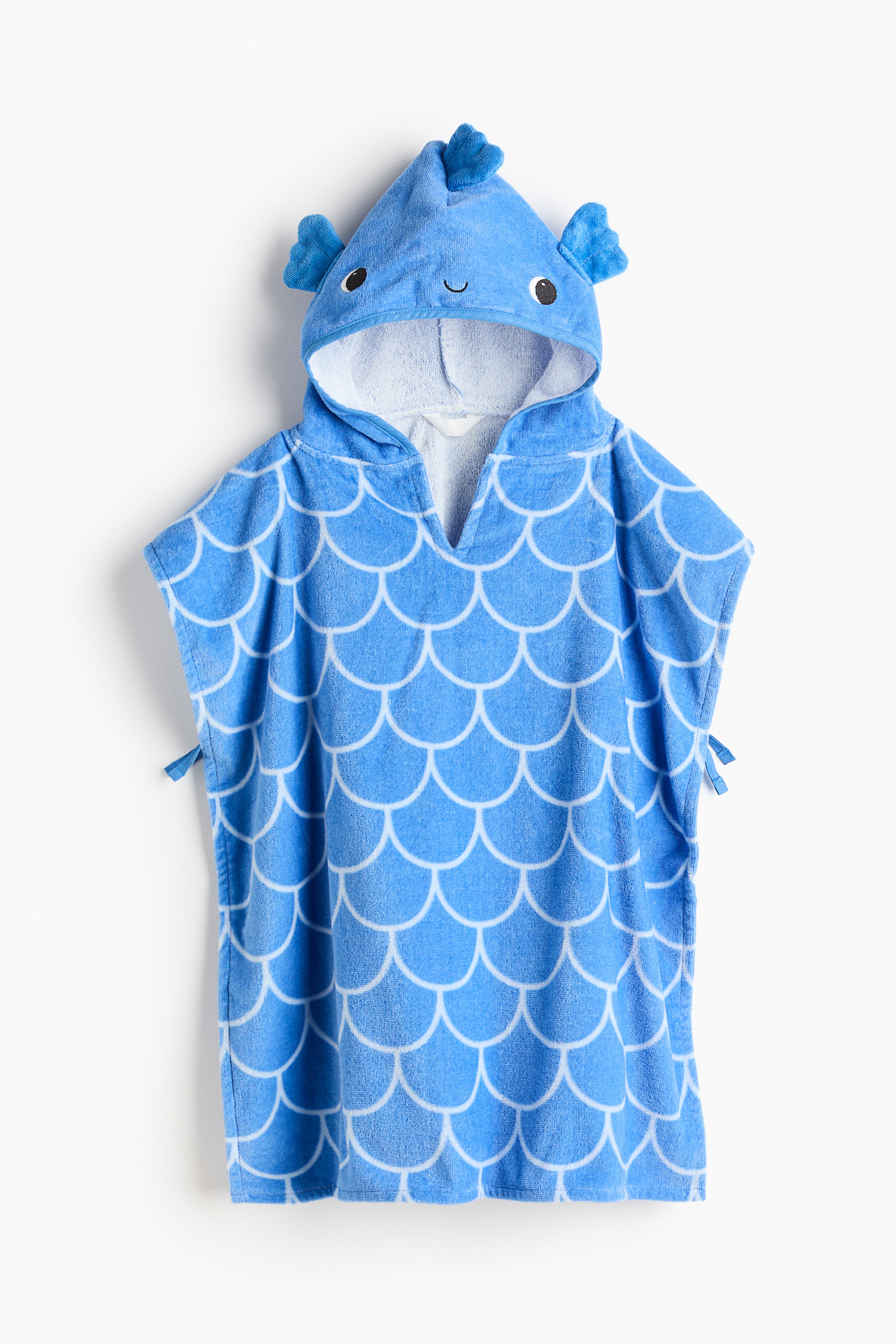 拡大画像を表示: 正面向きのタオルケープは、白い鱗模様の入った青いフード付きポンチョです。フードは魚の頭を模したデザインで、刺繍の目や口、小さなヒレがあしらわれています。ポンチョの両側には小さな青い紐が見えます。