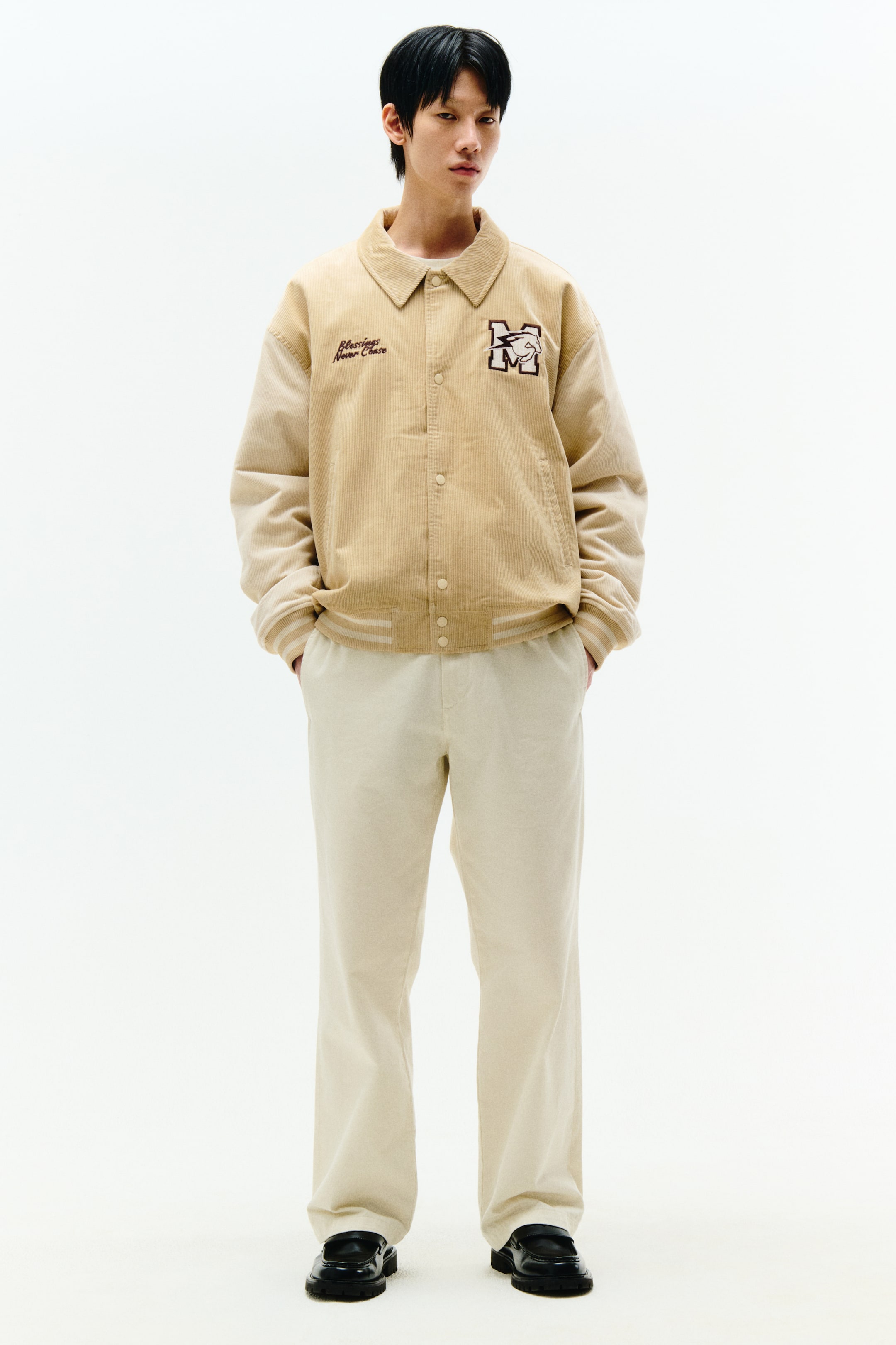 拡大画像を表示: ベージュのバーシティジャケットは前を向いており、コーデュロイのボディと、前にスナップボタン留めのあるより淡いベージュのコントラストスリーブが特徴です。左胸には刺繍されたテキストを表示し、右胸には動物デザインの大きな「M」ロゴがあり、リブ付きのストライプカフスとへムが付属しています。