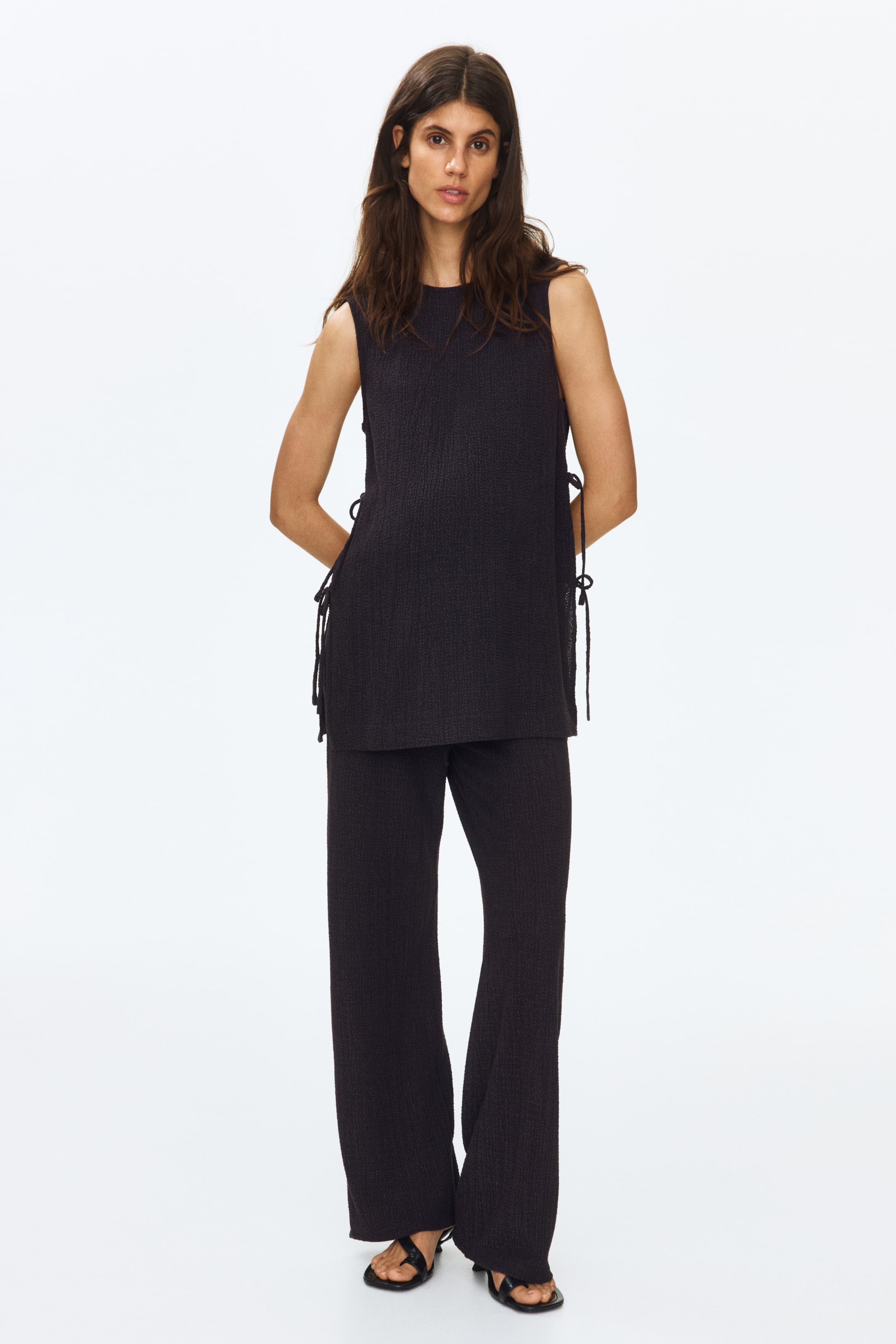 View larger image: A set in dark crinkled jersey, consisting of a long, sleeveless top and straight-leg trousers, is being worn by the model. The top features a round neckline and open sides with two sets of narrow ties and a side panel underneath, while the trousers have a low drawstring waist. The model is also wearing black sandals.